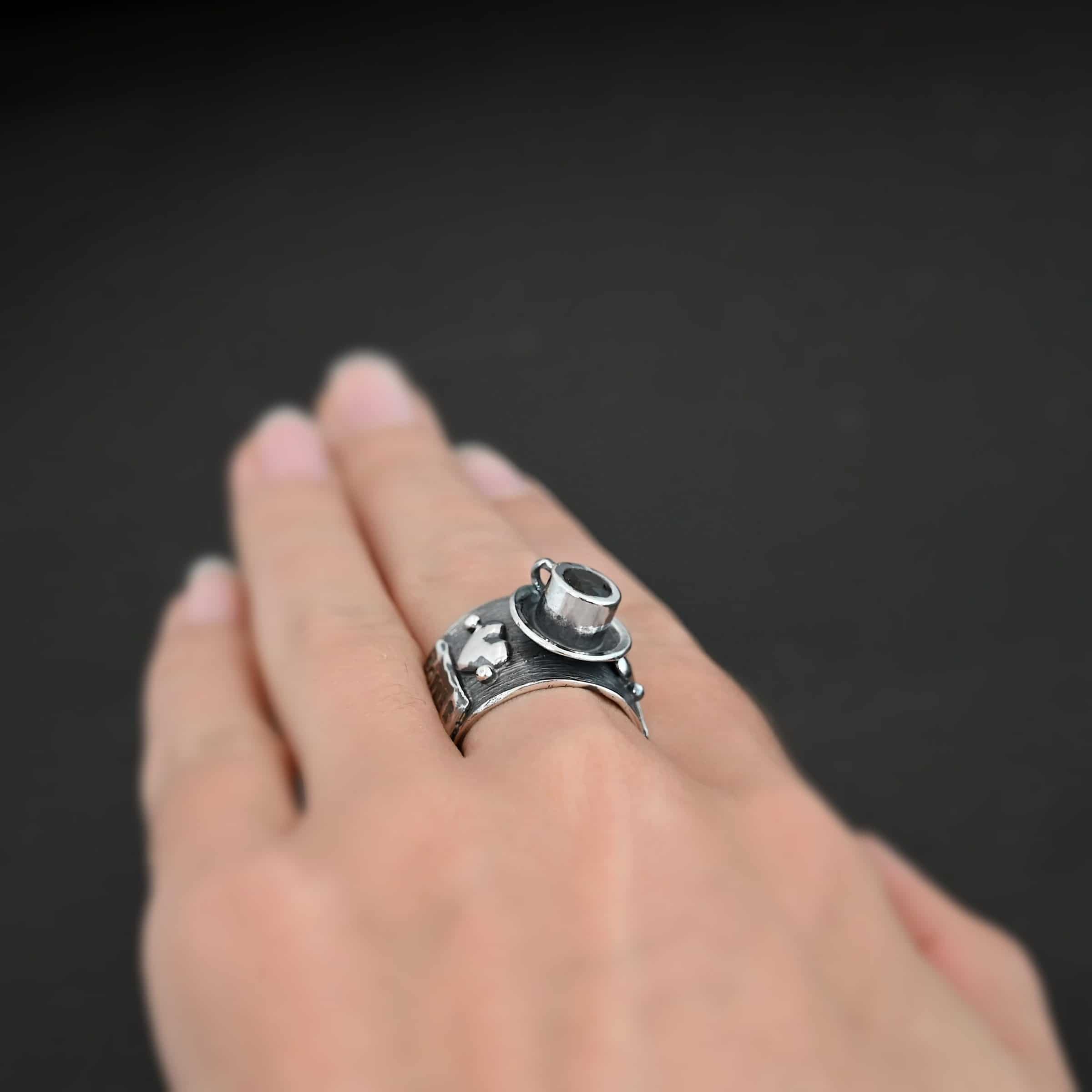 True Addiction Coffee Ring Photo of the "True Addiction Coffee Ring." Made out of sterling silver and smokey topaz. The side of the ring has "Coffee" stamped into it. Above that is a heart with a silver dot on each side of it. Above this, in the center of the ring, is a saucer with a cup and a handle, and a smokey topaz in the center of the cup representing the coffee. Ring is worn on a models middle finger to demonstrate size.