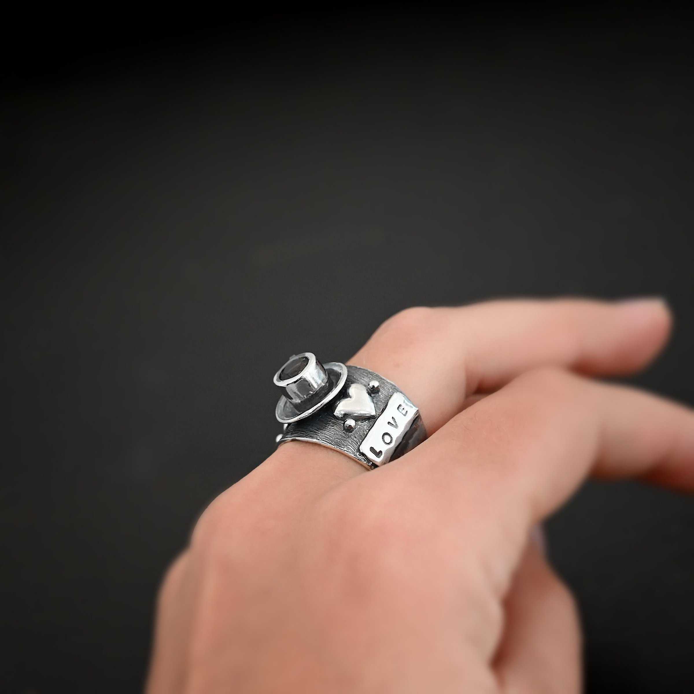 True Addiction Coffee Ring Photo of the "True Addiction Coffee Ring." Made out of sterling silver and smokey topaz. The side of the ring has "Love" stamped into it. Above that is a heart with a silver dot on each side of it. Above this, in the center of the ring, is a saucer with a cup and a handle, and a smokey topaz in the center of the cup representing the coffee. Ring is worn on a models middle finger to demonstrate size.