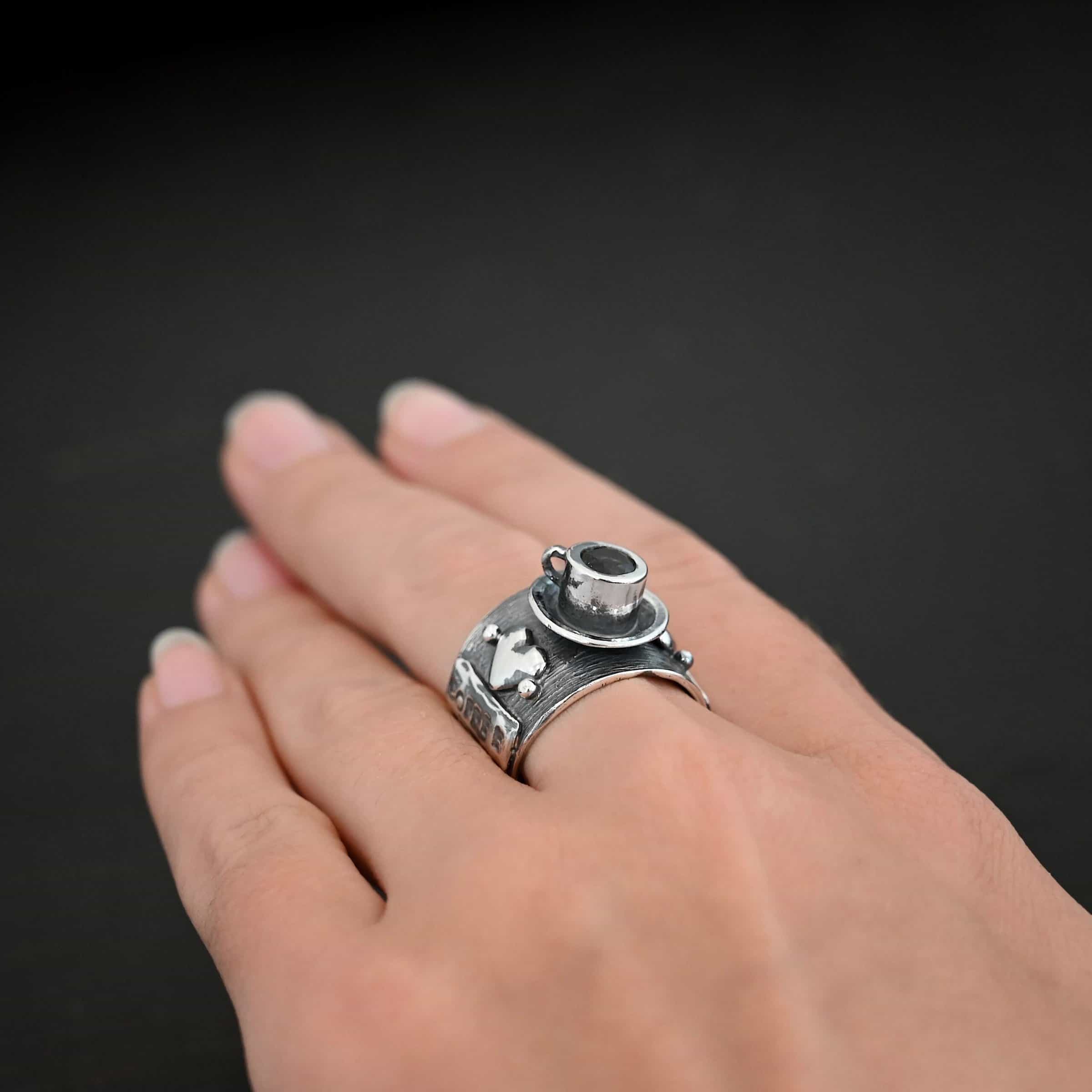 True Addiction Coffee Ring Photo of the "True Addiction Coffee Ring." Made out of sterling silver and smokey topaz. The side of the ring has "Coffee" stamped into it. Above that is a heart with a silver dot on each side of it. Above this, in the center of the ring, is a saucer with a cup and a handle, and a smokey topaz in the center of the cup representing the coffee. Ring is worn on a models middle finger to demonstrate size.