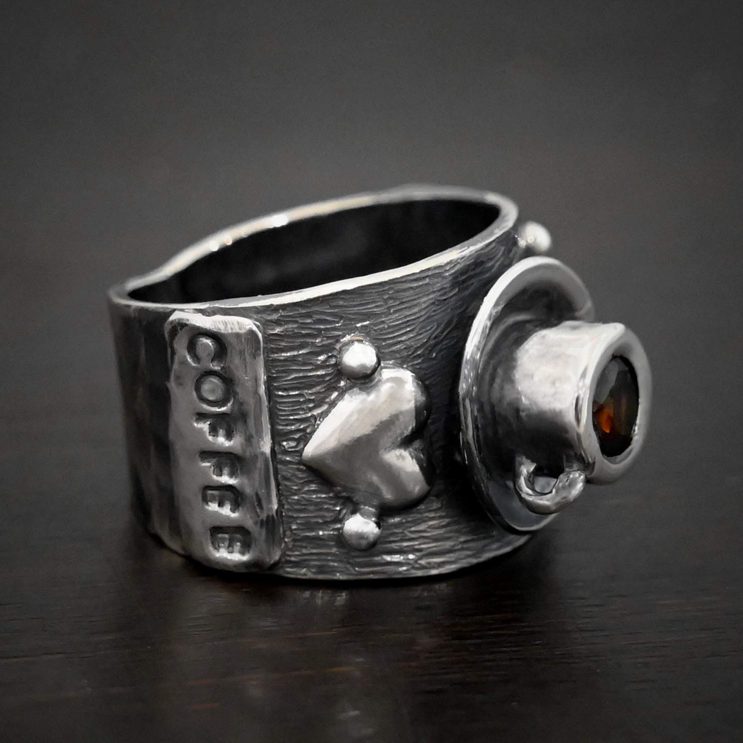 True Addiction Coffee Ring Photo of the "True Addiction Coffee Ring." Made out of sterling silver and smokey topaz. The side of the ring has "Coffee" stamped into it. Above that is a heart with a silver dot on each side of it. Above this, in the center of the ring, is a saucer with a cup and a handle, and a smokey topaz in the center of the cup representing the coffee. Ring is sitting against a dark background.