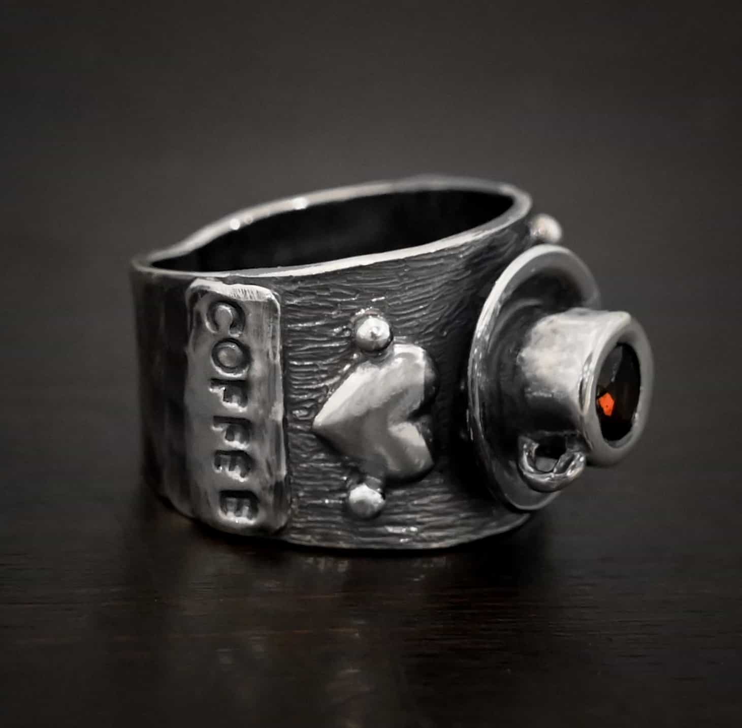 True Addiction Coffee Ring Photo of the "True Addiction Coffee Ring." Made out of sterling silver and smokey topaz. The side of the ring has "Coffee" stamped into it. Above that is a heart with a silver dot on each side of it. Above this, in the center of the ring, is a saucer with a cup and a handle, and a smokey topaz in the center of the cup representing the coffee. Ring is sitting against a dark background.