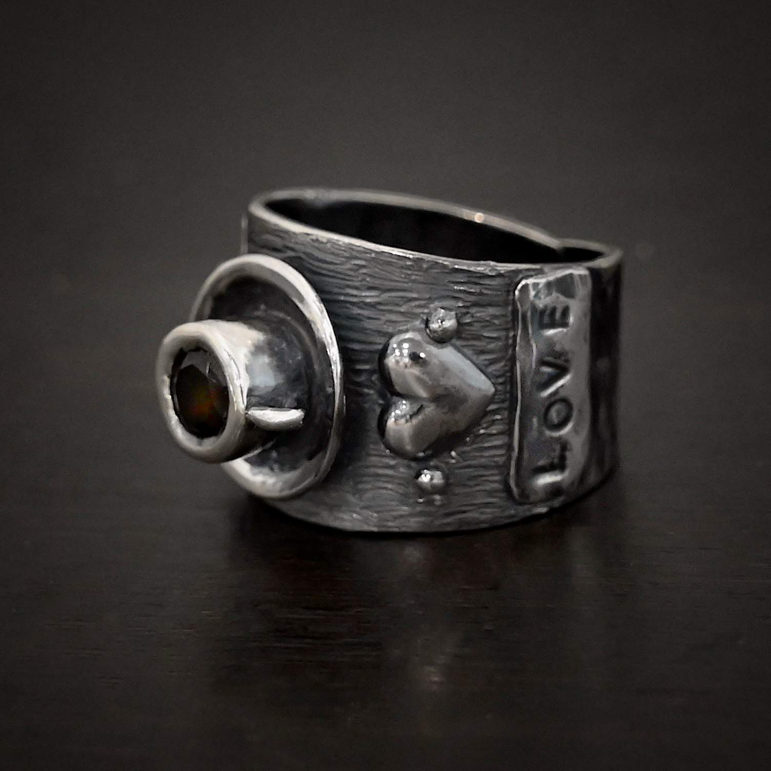 True Addiction Coffee Ring Photo of the "True Addiction Coffee Ring." Made out of sterling silver and smokey topaz. The side of the ring has "Love" stamped into it. Above that is a heart with a silver dot on each side of it. Above this, in the center of the ring, is a saucer with a cup and a handle, and a smokey topaz in the center of the cup representing the coffee. Ring is sitting against a dark background.