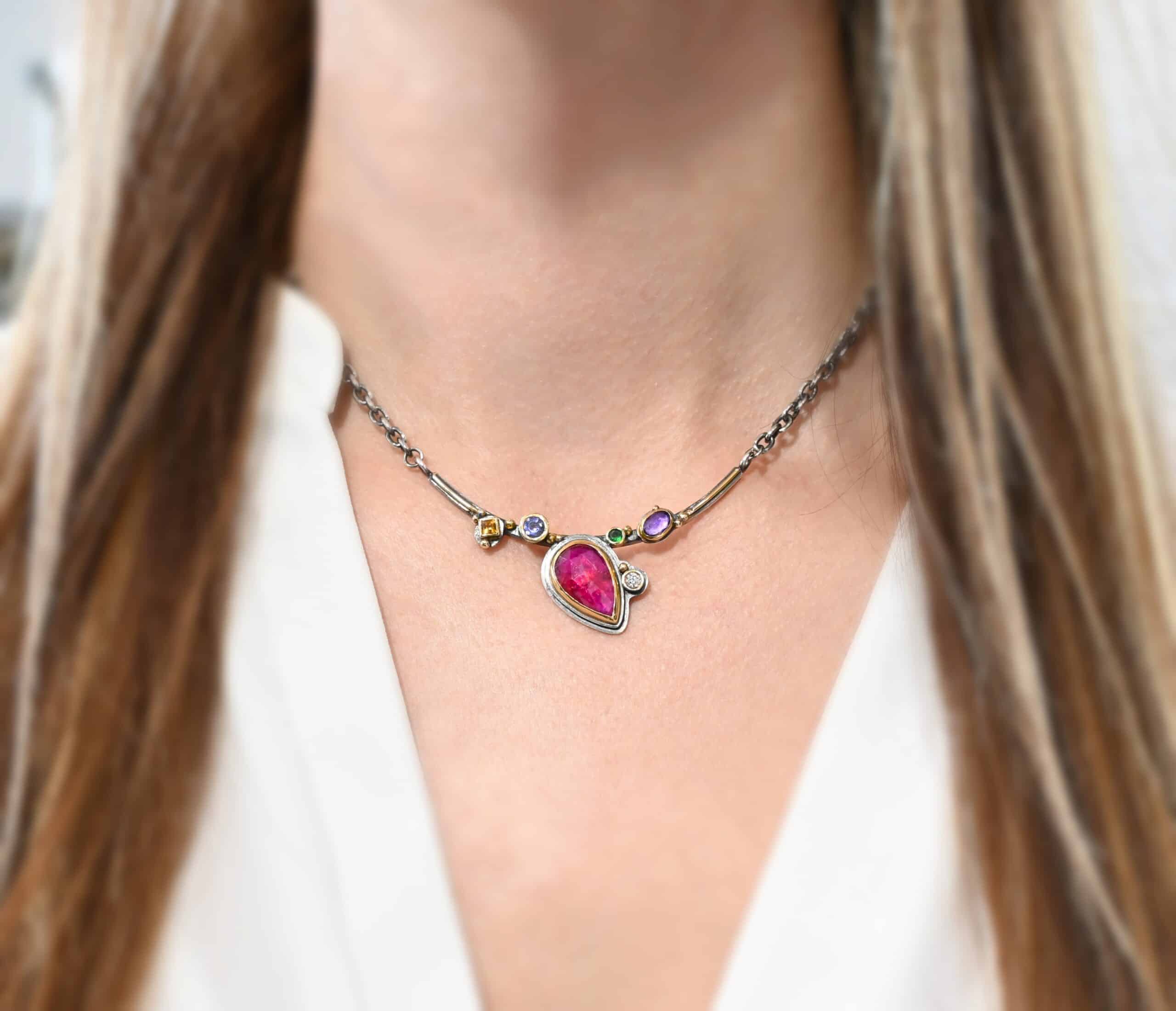 Photo of the "Cinderella" Necklace. It has a ruby in the center, pear shaped and set pointing to the bottom right. There is an amethyst and citrine on the left side and a tsavorite garnet and amethyst on the right side. There is a diamond on the bottom right side of the ruby. Each stone is set in gold and soldered to a curved bar. There are multiple gold balls scattered around the bar and stones. The bar has a thin line of gold going across it where the stones aren't. Necklace is worn by a model.