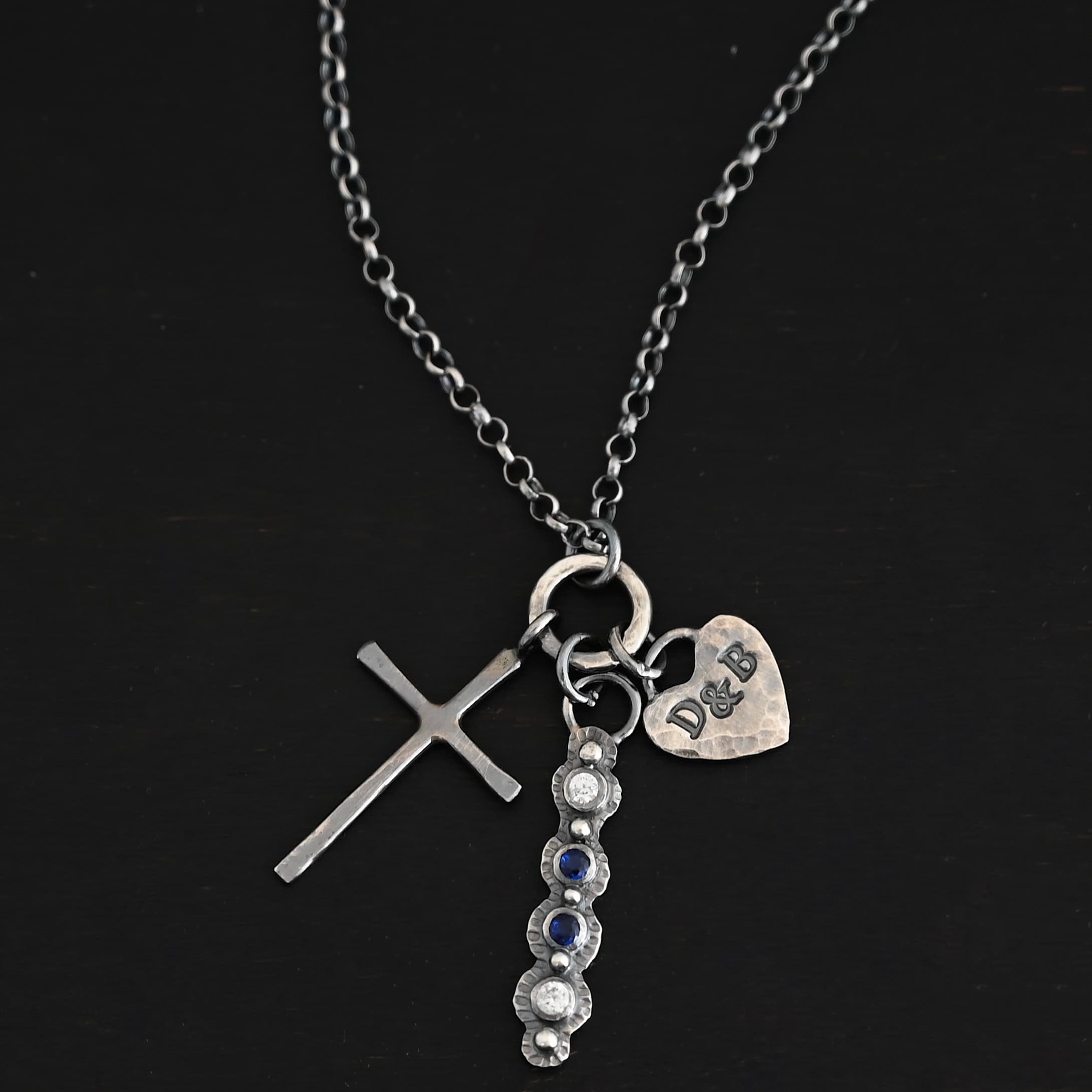 Photo of a custom "Mother's Necklace." Made out of sterling silver and four different birthstones. The top stone is clear, then there is two blue stones, and another clear one at the bottom. Each stone is a small circle, and the silver around them is hammered and oxidized. There are five silver balls on the necklace that go in a straight line. There is one on the top of the first stone, and each ball after that separates each stone. There is a loop at the top of the pendant with a jump ring through it that is attached to a larger hammered circle. There is a small cross charm on the left side of the pendant, and a heart charm on the right side of the pendant with the initials "D & B" stamped. Both are attached to the large circle as well. The large circle has a small jump ring attached to the top of it in which the chain goes through. Necklace is laying against a dark background.