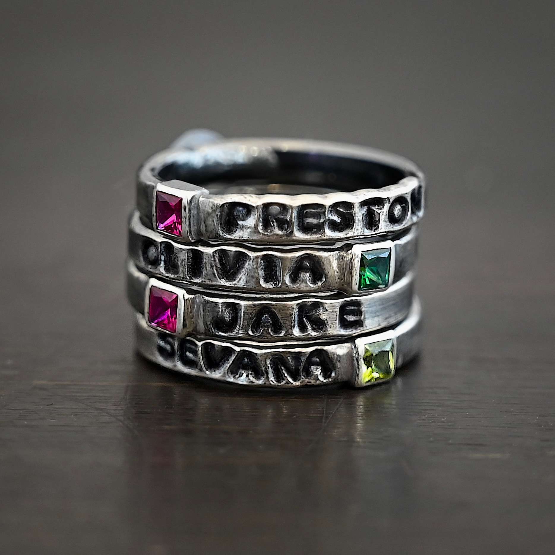 Photo of personalized birthstone stacker rings. The top ring has a square ruby with the name "Preston" stamped and colored in with black on the right side of the ruby. The second ring has the name "Olivia" on it with a square emerald on the right side of the ring. The third ring has the name "Jake" on it with the square ruby on the left side of the name. The final ring has the name "Sevana" on it with a square peridot on the right side of the name. Rings are sitting on top of each other against a black background.