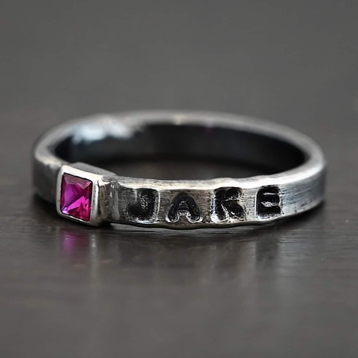 Photo of personalized birthstone stacker rings. The center ring is silver and has a square ruby in it. On the right side of the ruby, the name "Jake" is stamped and colored in with black. Ring is laying against a black background.