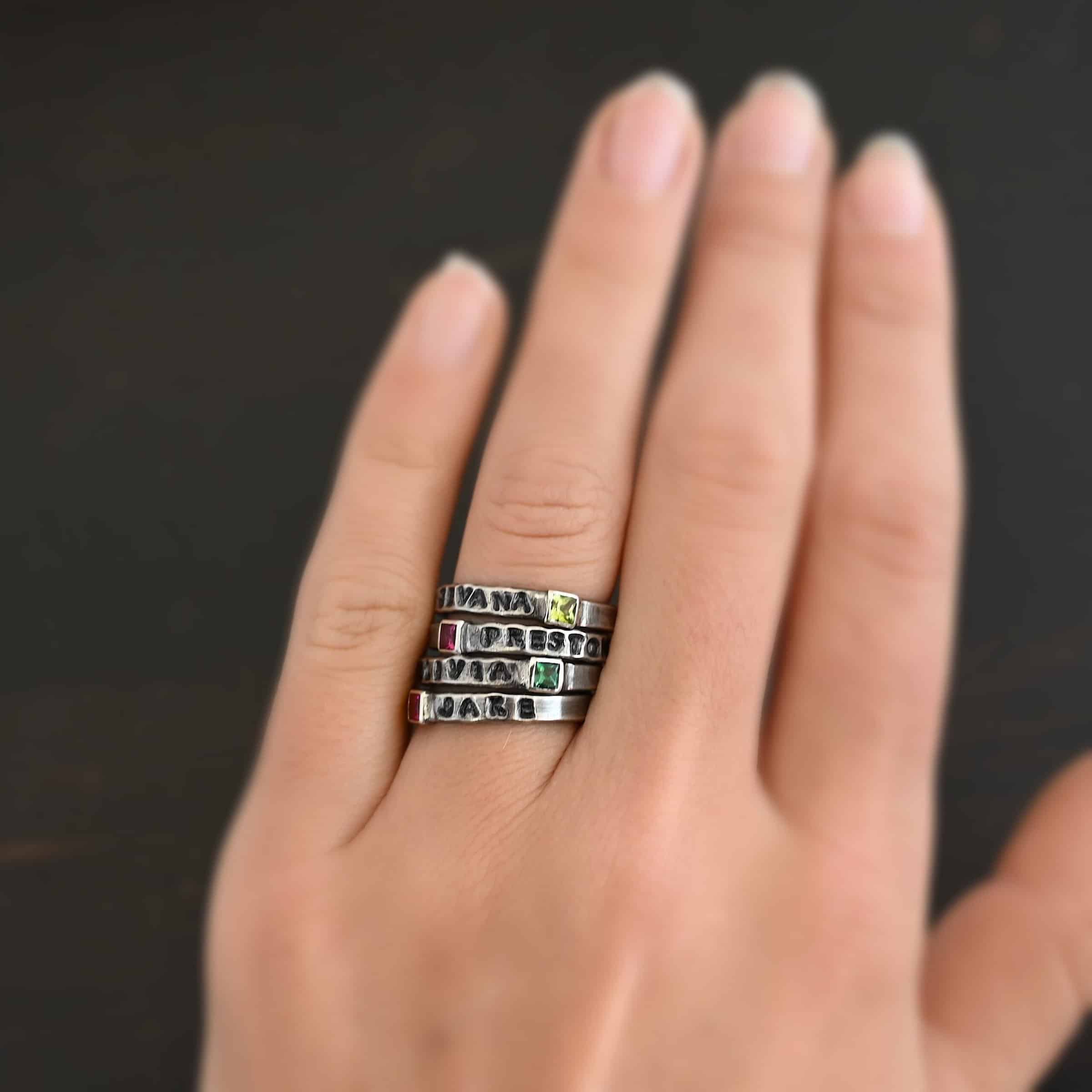 Photo of personalized birthstone stacker rings. The top ring has the name "Sevana" on it with a square peridot on the right side of the name. The second ring has a square ruby with the name "Preston" stamped and colored in with black on the right side of the ruby. The third ring has the name "Olivia" on it with a square emerald on the right side of the ring. The final ring has the name "Jake" on it with the square ruby on the left side of the name. Rings are worn on a models engagement ring finger to demonstrate how they look together.