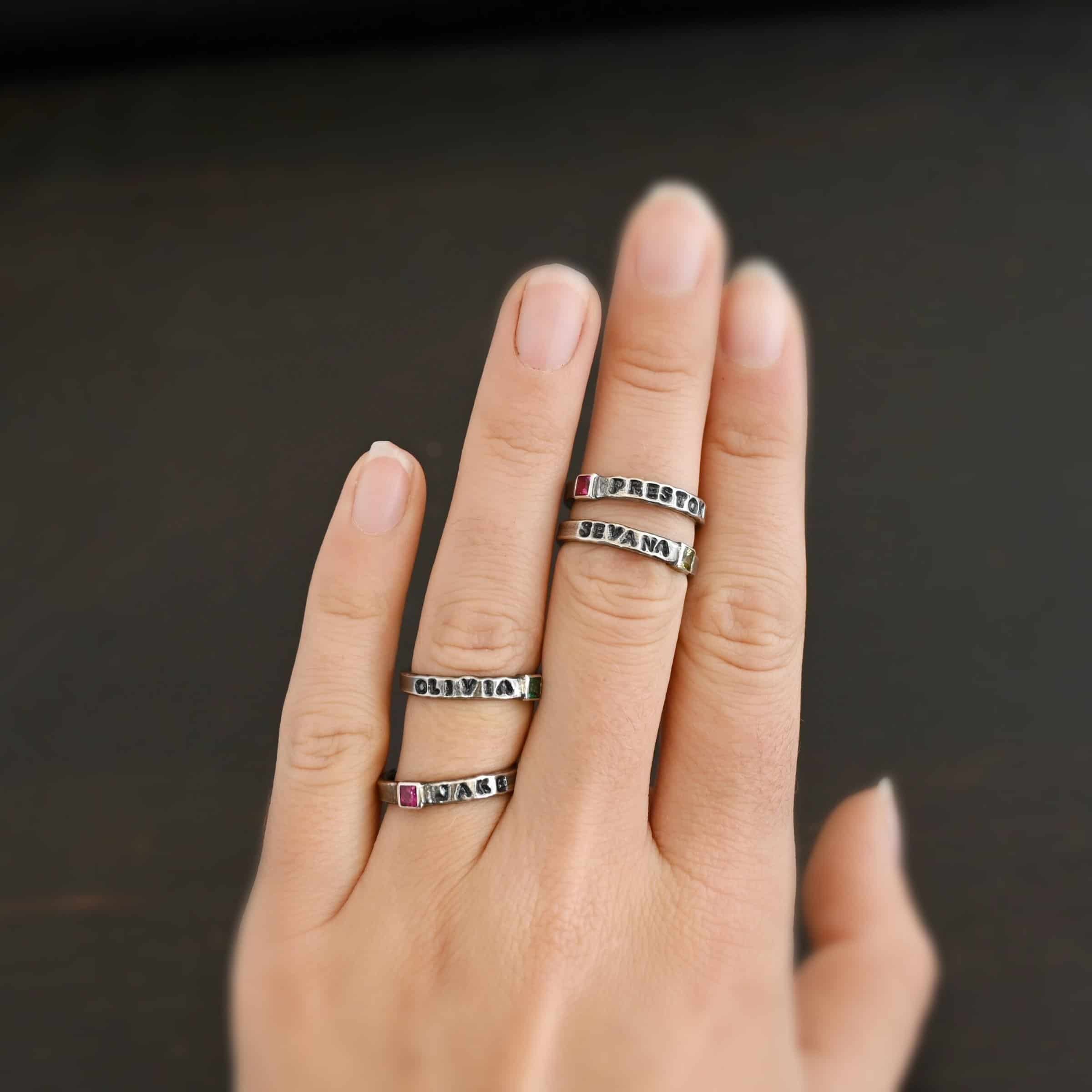 Photo of personalized birthstone stacker rings. "Olivia" and "Jake" are on the engagement ring finger and "Preston" and "Sevana" are on the middle finger. The top ring on the engagement ring finger has the name "Olivia" on it with a square emerald on the right side of the ring. The second ring on that finger has the name "Jake" on it with the square ruby on the left side of the name. The top ring on the middle finger has a square ruby with the name "Preston" stamped and colored in with black on the right side of the ruby. The bottom ring has the name "Sevana" on it with a square peridot on the right side of the name. Rings are worn on a models engagement ring finger and middle finger to demonstrate how they look together.