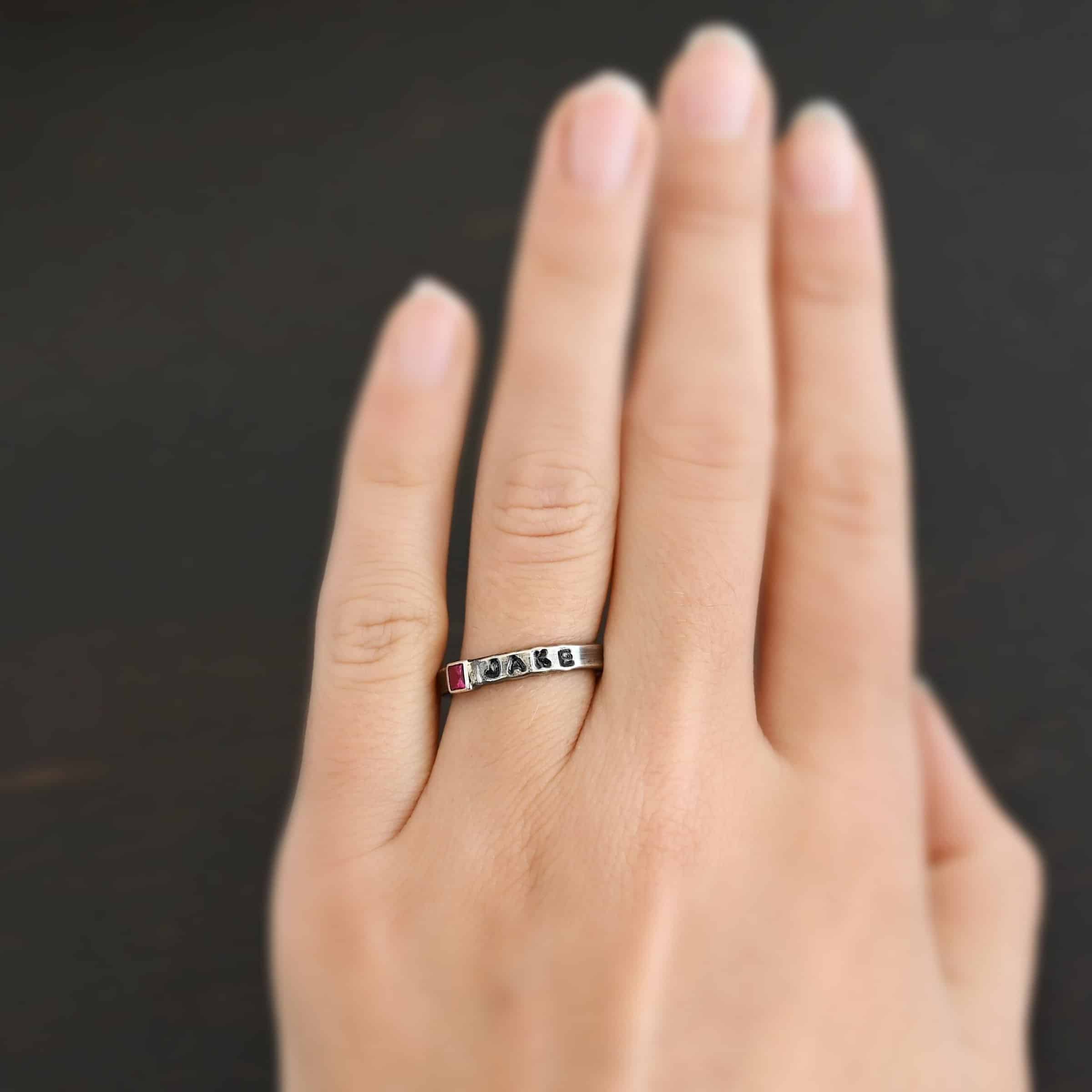 Photo of personalized birthstone stacker rings. The center ring is silver and has a square ruby in it. On the right side of the ruby, the name "Jake" is stamped and colored in with black. Ring is worn on a models engagement ring finger to demonstrate size.