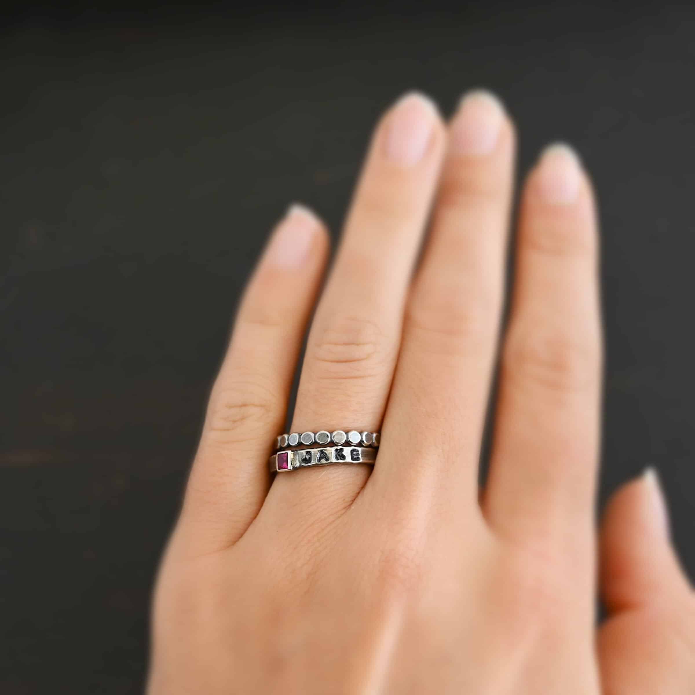 Photo of personalized birthstone stacker rings. The center ring is silver and has a square ruby in it. On the right side of the ruby, the name "Jake" is stamped and colored in with black. Ring is worn on a models engagement ring finger with another silver stacker ring that is made of small hammered silver dots that are soldered together to demonstrate size.