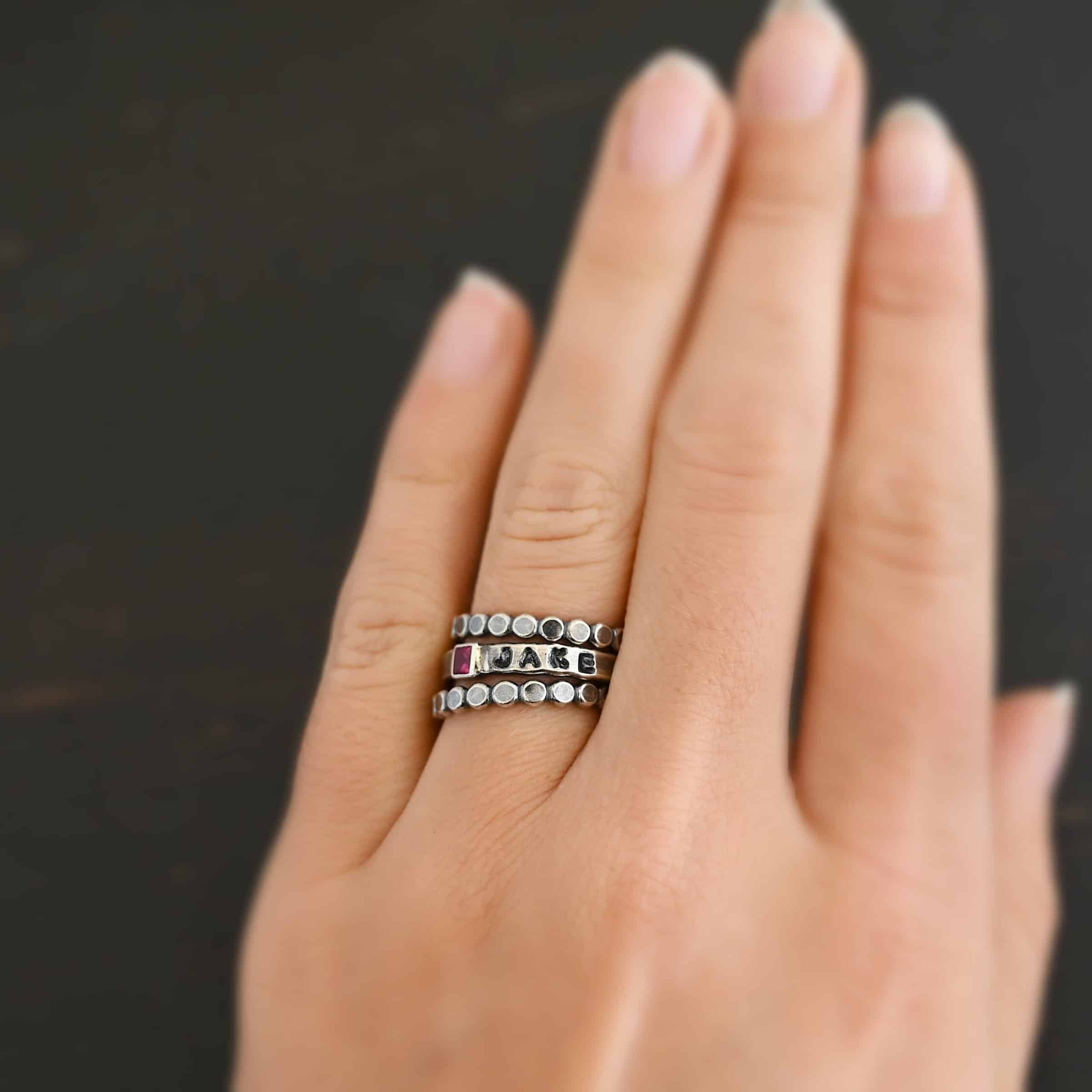 Photo of personalized birthstone stacker rings. The center ring is silver and has a square ruby in it. On the right side of the ruby, the name "Jake" is stamped and colored in with black. There is a ring above and below the middle ring. Both are sterling silver, and are small hammered circles that are soldered together. Rings are worn on a models ring finger to demonstrate how the rings look together.