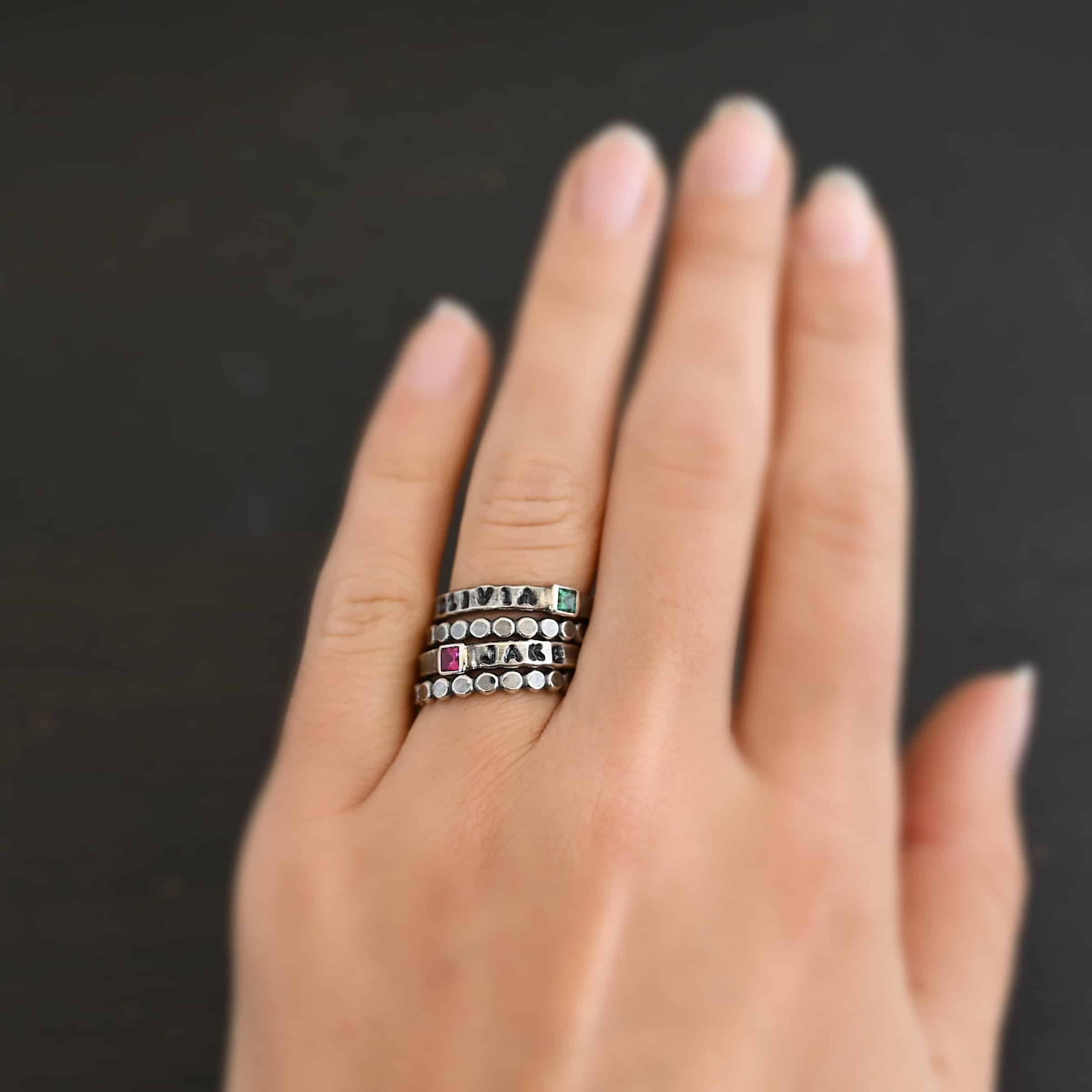 Photo of personalized birthstone stacker rings. "Olivia" and "Jake" are worn together with stacker rings that are small hammered dots soldered together. The top ring on the engagement ring finger has the name "Olivia" on it with a square emerald on the right side of the ring. Then there is a stacker ring. The second ring on that finger has the name "Jake" on it with the square ruby on the left side of the name. There is a stacker ring beneath that ring. Ring is worn on an engagement ring finger to demonstrate size and how they look together.
