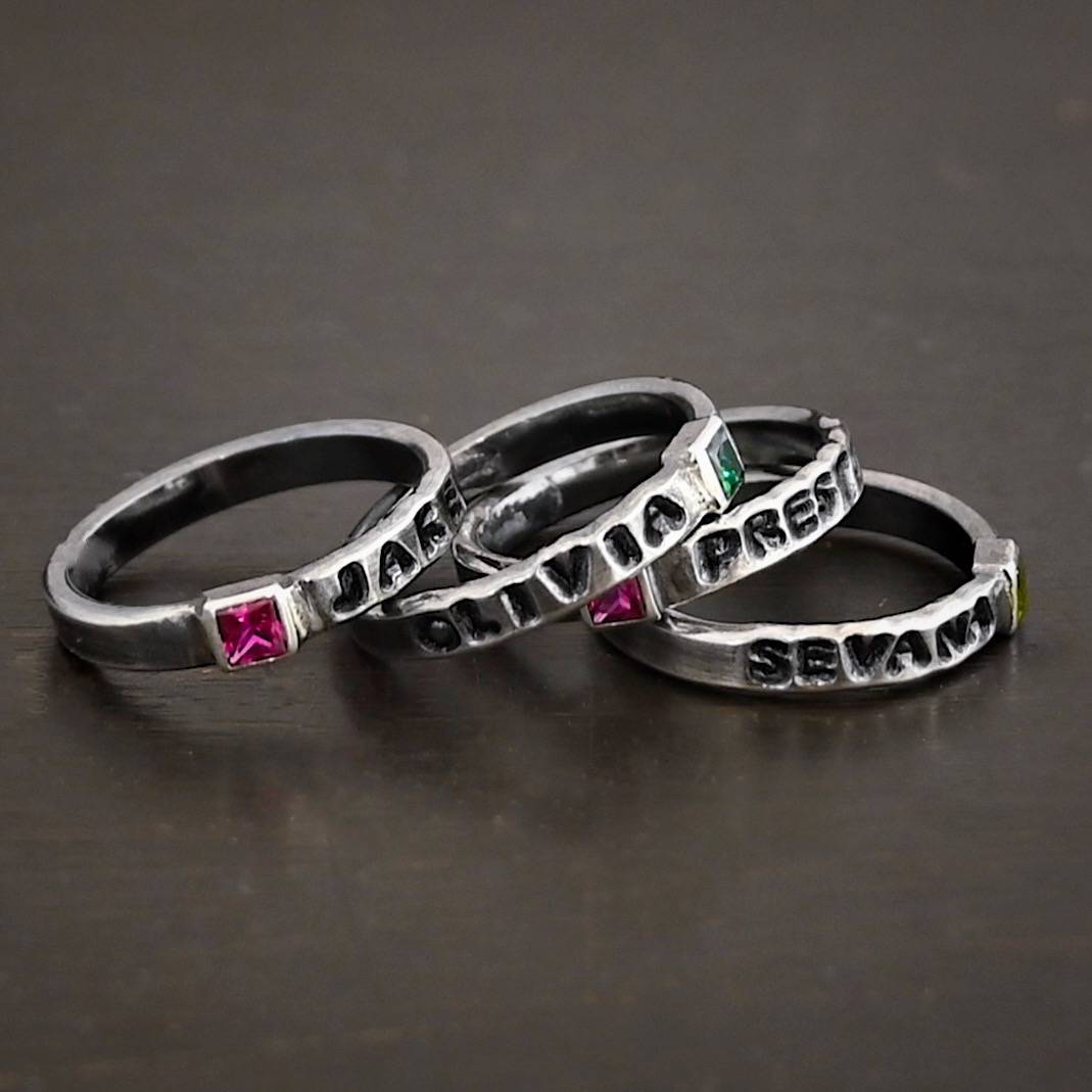 Photo of personalized birthstone stacker rings. The top ring has the name "Jake" on it with the square ruby on the left side of the name. The second ring has the name "Olivia" on it with a square emerald on the right side of the ring. The third ring has a square ruby with the name "Preston" stamped and colored in with black on the right side of the ruby. The final ring has the name "Sevana" on it with a square peridot on the right side of the name. Rings are stacked from left to right on top of each other against a black background.