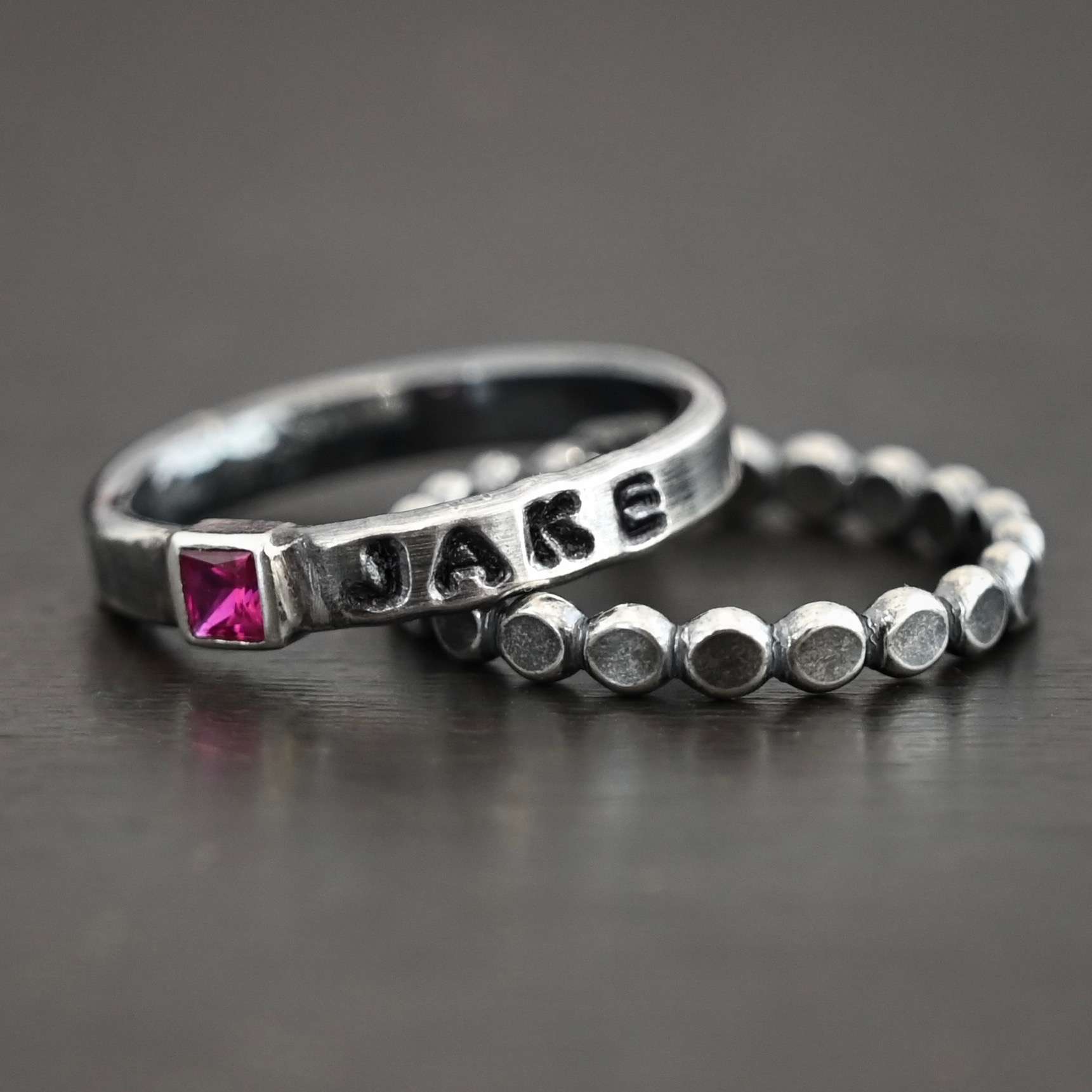 Photo of personalized birthstone stacker rings. The center ring is silver and has a square ruby in it. On the right side of the ruby, the name "Jake" is stamped and colored in with black. Ring 9s sitting against a black background with another silver stacker ring that is made of small hammered silver dots that are soldered together.