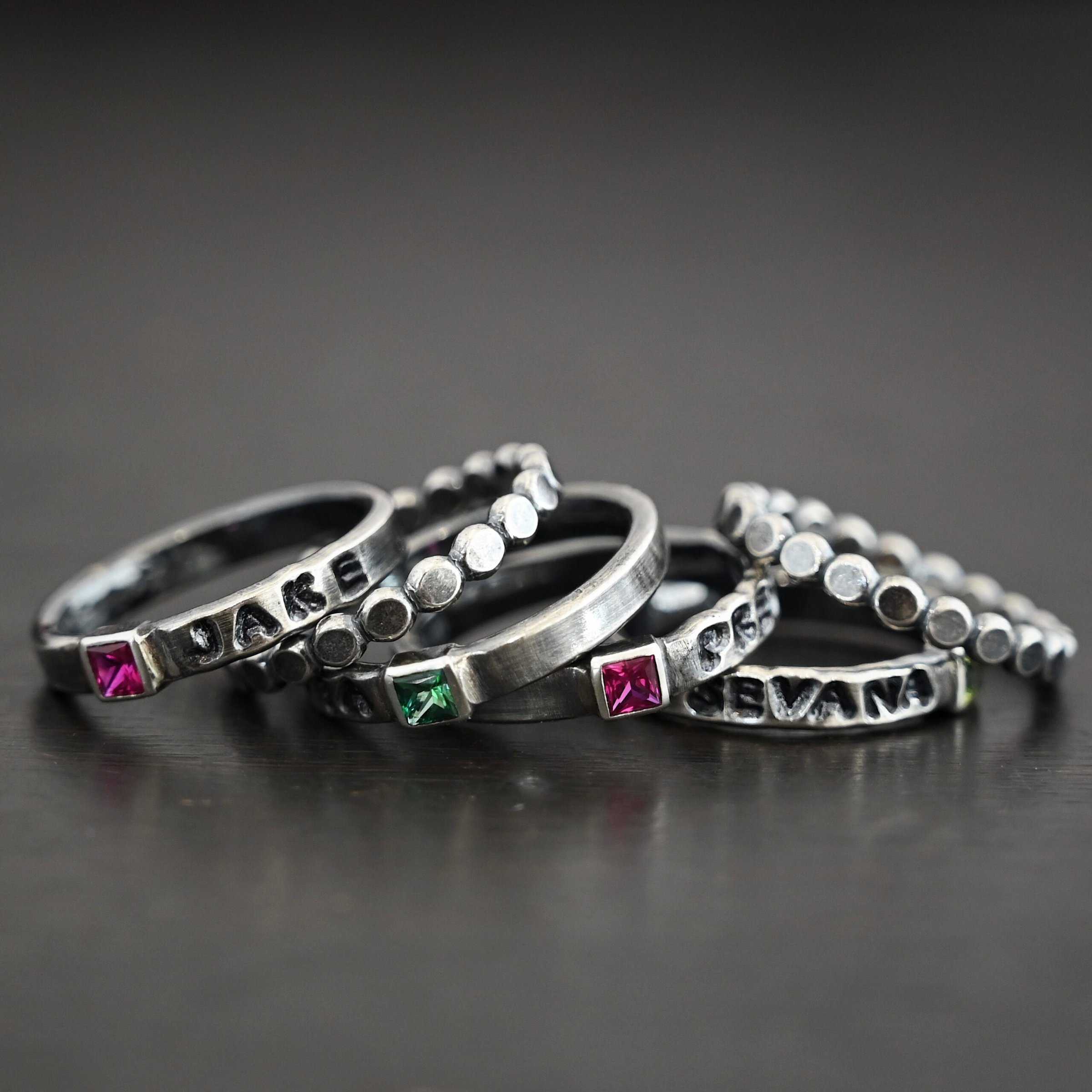 Photo of personalized birthstone stacker rings. The top ring has the name "Jake" on it with the square ruby on the left side of the name. Then there is a silver ring made out of hammered dots soldered together. The second ring has the name "Olivia" on it with a square emerald on the right side of the ring. The third ring has a square ruby with the name "Preston" stamped and colored in with black on the right side of the ruby. The final ring has the name "Sevana" on it with a square peridot on the right side of the name. Then there is another stacker ring made out of hammered soldered dots. Rings are stacked from left to right on top of each other against a black background.