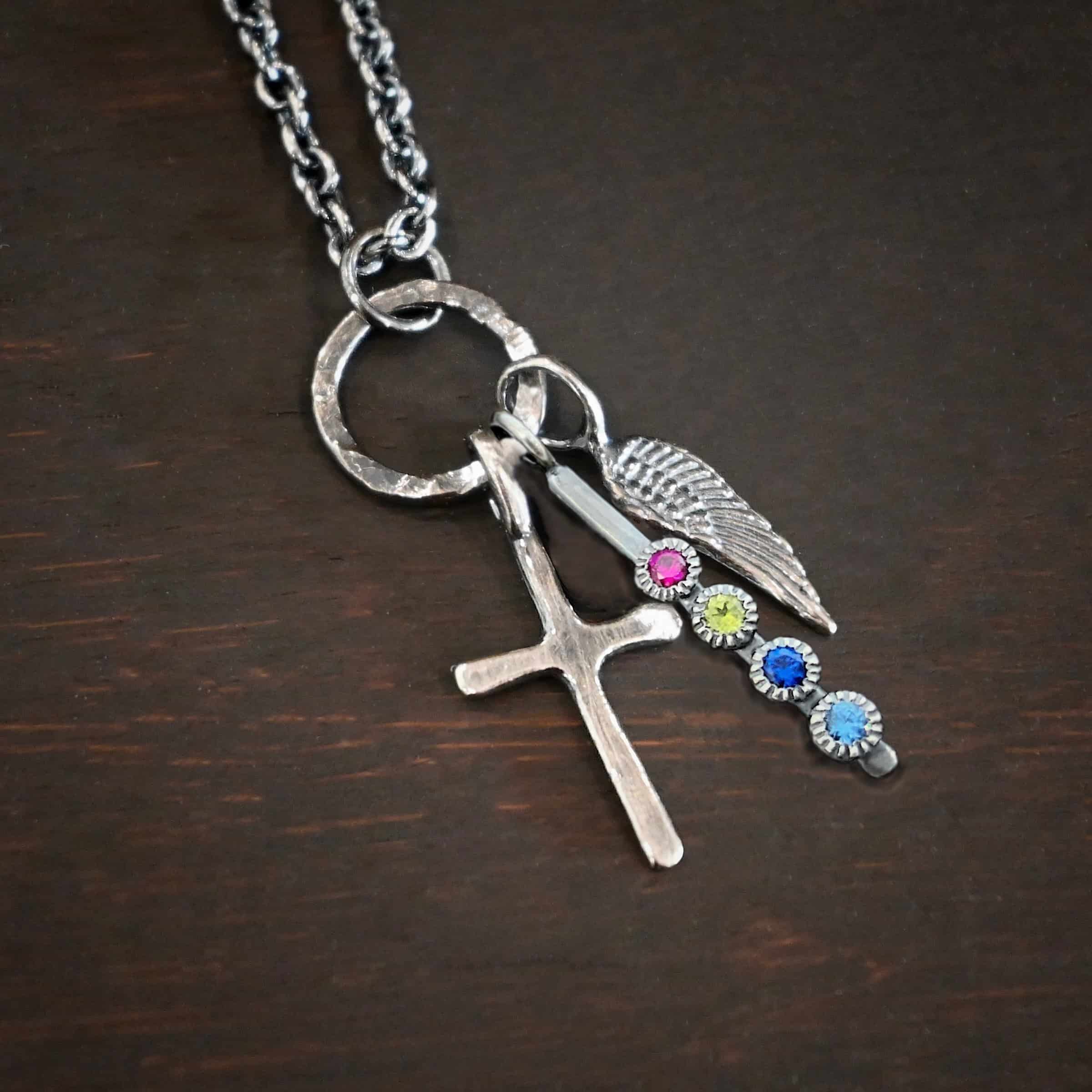 Photo of a cross and angel wing necklace. Made out of oxidized sterling silver. There is a small sized cross that is flat and smooth. It has a loop soldered to the top of it. There is a small sized angel wing pendant next to it, also with a soldered loop on the top. There is a small, long rectangle with four birthstones on it and a soldered loop on the top. The birthstones are ruby, peridot, sapphire, and aquamarine. There is a chain that goes through the top of the necklace that is silver and oxidized. Necklace is lying against a dark background.