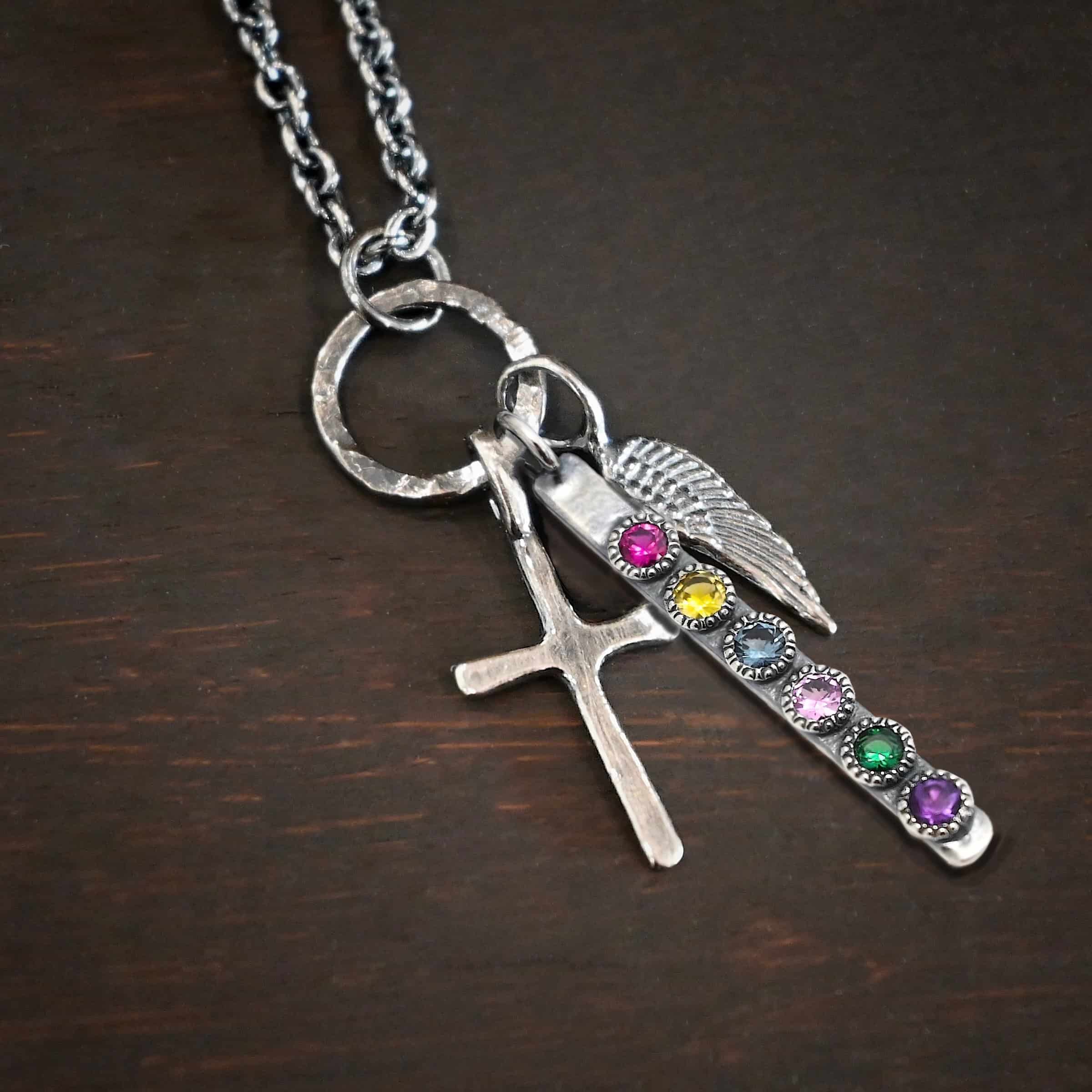 Photo of a cross and angel wing necklace. Made out of oxidized sterling silver. There is a small sized cross that is flat and smooth. It has a loop soldered to the top of it. There is a small sized angel wing pendant next to it, also with a soldered loop on the top. There is a small, long rectangle with six birthstones on it and a soldered loop on the top. The birthstones are ruby, citrine, alexandrite, pink zircon, emerald, and amethyst. There is a chain that goes through the top of the necklace that is silver and oxidized. Necklace is lying against a dark background.