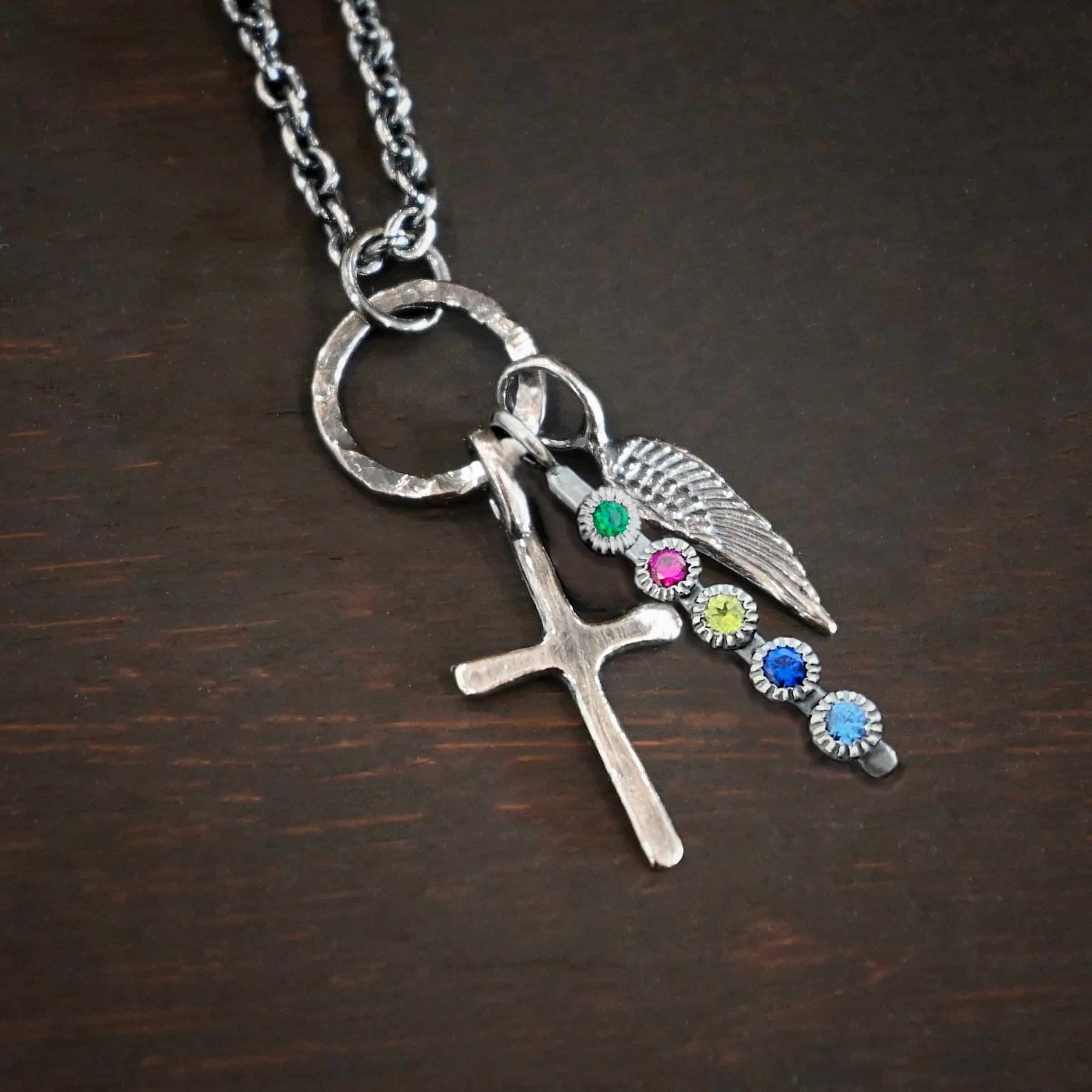 Photo of a cross and angel wing necklace. Made out of oxidized sterling silver. There is a small sized cross that is flat and smooth. It has a loop soldered to the top of it. There is a small sized angel wing pendant next to it, also with a soldered loop on the top. There is a small, long rectangle with five birthstones on it and a soldered loop on the top. The birthstones are an emerald, ruby, peridot, sapphire, and aquamarine. There is a chain that goes through the top of the necklace that is silver and oxidized. Necklace is lying against a dark background.