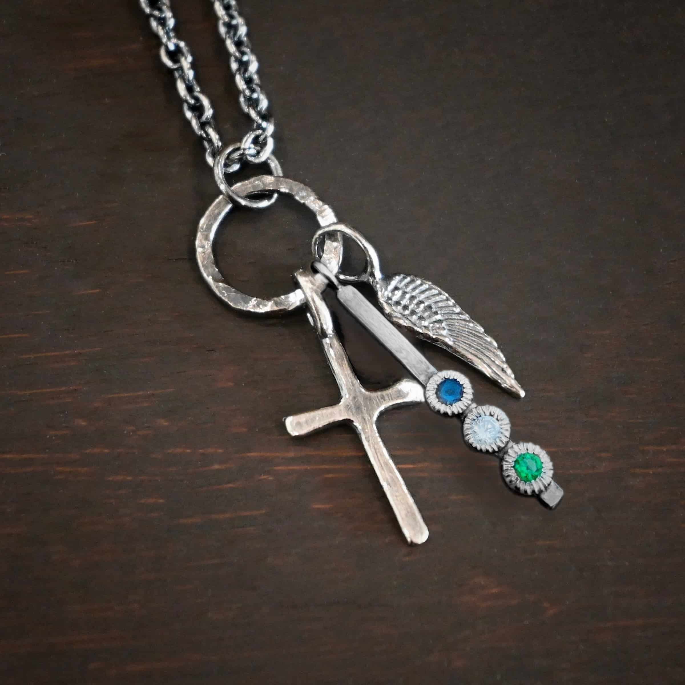 Photo of a cross and angel wing necklace. Made out of oxidized sterling silver. There is a small sized cross that is flat and smooth. It has a loop soldered to the top of it. There is a small sized angel wing pendant next to it, also with a soldered loop on the top. There is a small, long rectangle with three birthstones on it and a soldered loop on the top. The birthstones are sapphire, aquamarine, and emerald. There is a chain that goes through the top of the necklace that is silver and oxidized. Necklace is lying against a dark background.