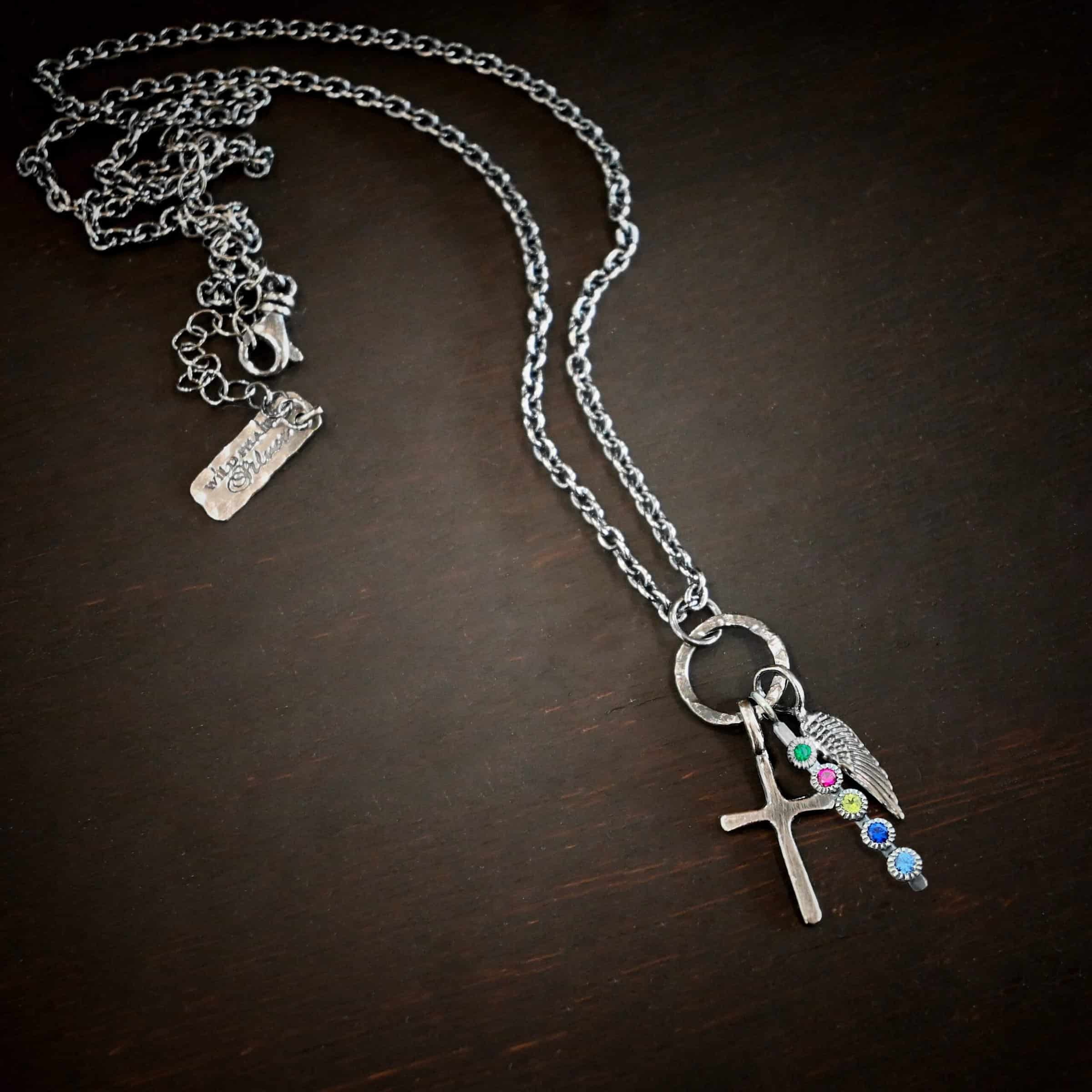 Photo of a cross and angel wing necklace. Made out of oxidized sterling silver. There is a small sized cross that is flat and smooth. It has a loop soldered to the top of it. There is a small sized angel wing pendant next to it, also with a soldered loop on the top. There is a small, long rectangle with five birthstones on it and a soldered loop on the top. The birthstones are an emerald, ruby, peridot, sapphire, and aquamarine. There is a chain that goes through the top of the necklace that is silver and oxidized. Necklace is lying against a dark background.