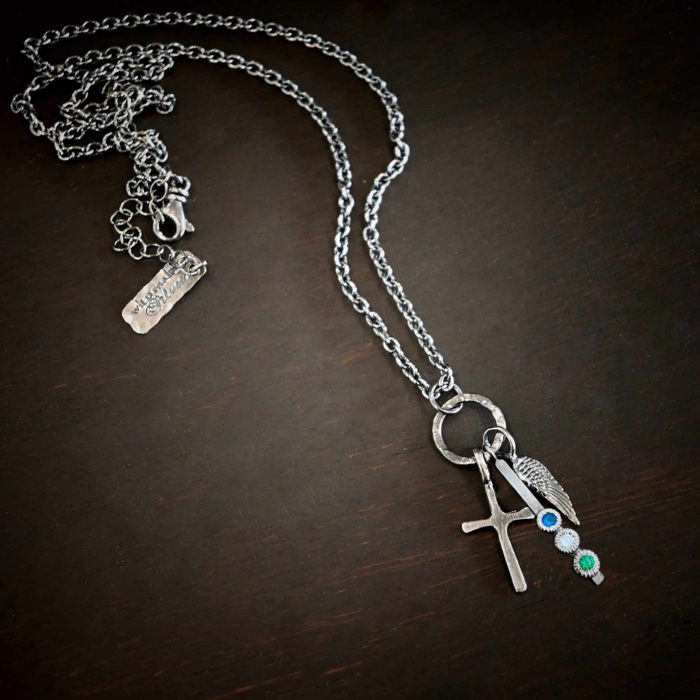 Photo of a cross and angel wing necklace. Made out of oxidized sterling silver. There is a small sized cross that is flat and smooth. It has a loop soldered to the top of it. There is a small sized angel wing pendant next to it, also with a soldered loop on the top. There is a small, long rectangle with three birthstones on it and a soldered loop on the top. The birthstones are sapphire, aquamarine, and emerald. There is a chain that goes through the top of the necklace that is silver and oxidized. Necklace is lying against a dark background.
