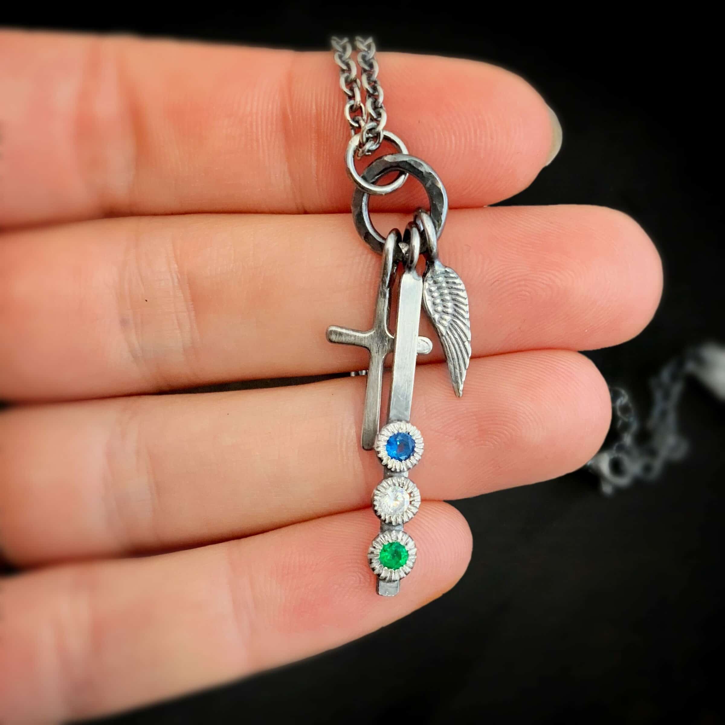 Photo of a cross and angel wing necklace. Made out of oxidized sterling silver. There is a small sized cross that is flat and smooth. It has a loop soldered to the top of it. There is a small sized angel wing pendant next to it, also with a soldered loop on the top. There is a small, long rectangle with three birthstones on it and a soldered loop on the top. The birthstones are sapphire, aquamarine, and emerald. There is a chain that goes through the top of the necklace that is silver and oxidized. Necklace is lying against the fingertips of a hand.