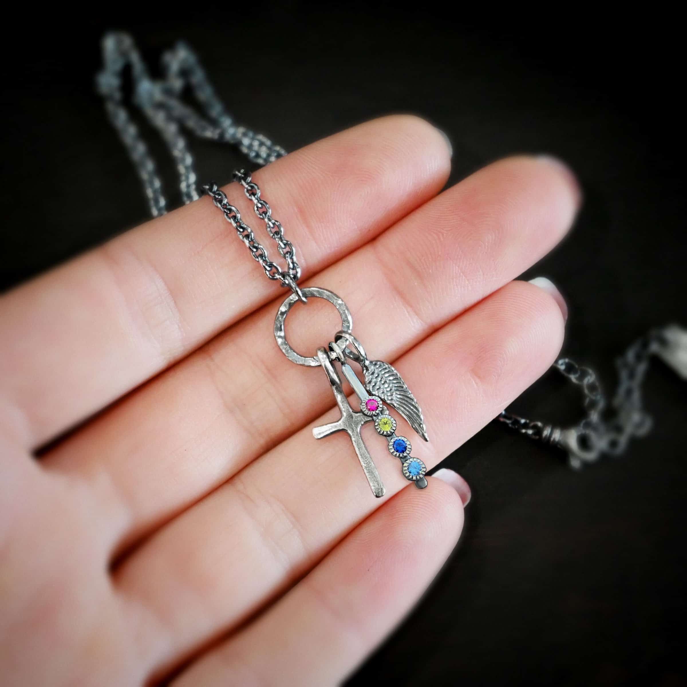 Photo of a cross and angel wing necklace. Made out of oxidized sterling silver. There is a small sized cross that is flat and smooth. It has a loop soldered to the top of it. There is a small sized angel wing pendant next to it, also with a soldered loop on the top. There is a small, long rectangle with four birthstones on it and a soldered loop on the top. The birthstones are ruby, peridot, sapphire, and aquamarine. There is a chain that goes through the top of the necklace that is silver and oxidized. Necklace is lying against the fingertips of a hand.