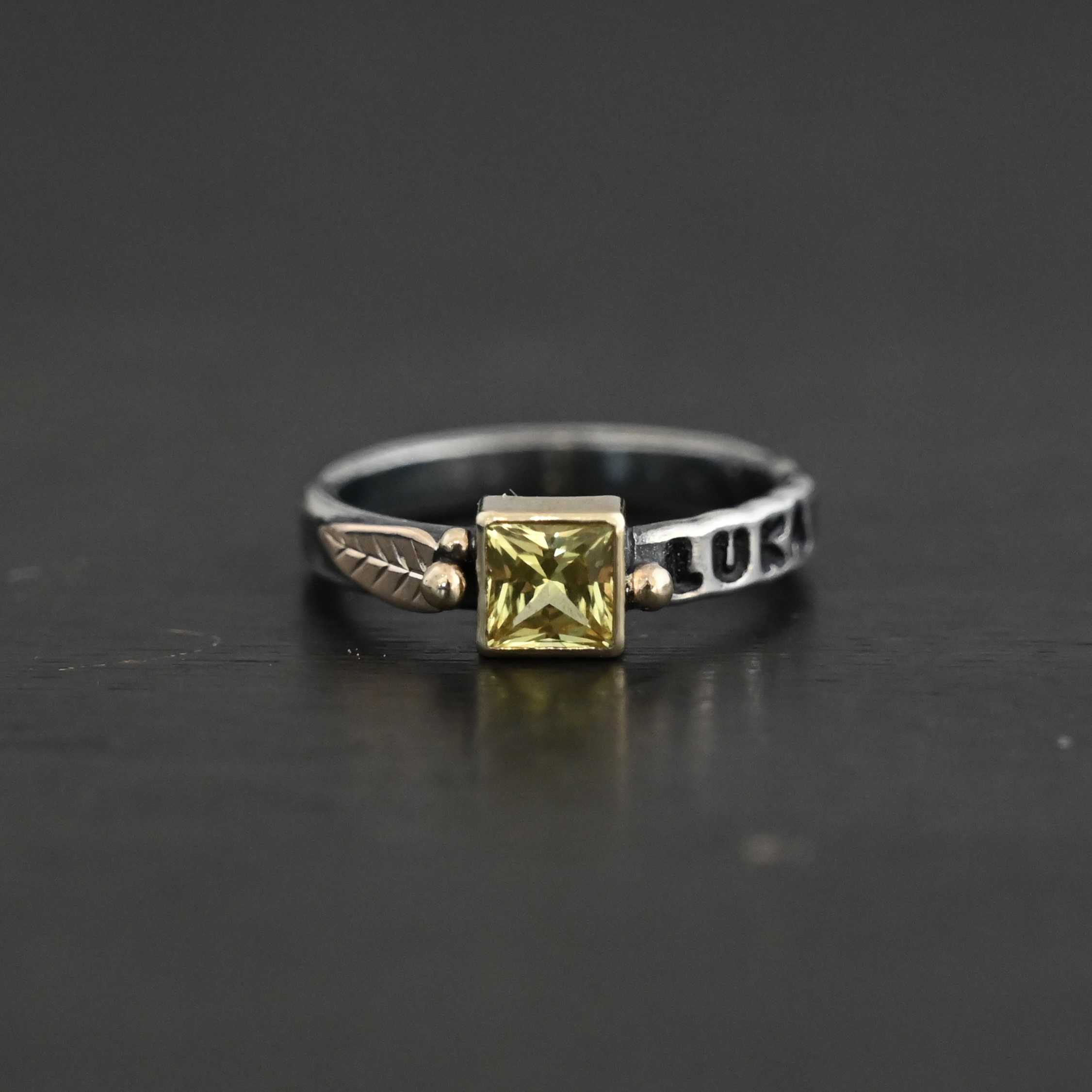 Photo of a personalized birthstone stacker. Ring is a silver band with a square citrine in the center of it. The citrine is set in gold. There is a gold dot on the right side of the stone setting, and two on the left side. There is also a gold leaf on the left side of the stone setting. The name "Luka" is stamped and colored in with black on the right side of the ring. Ring is sitting against a black background.