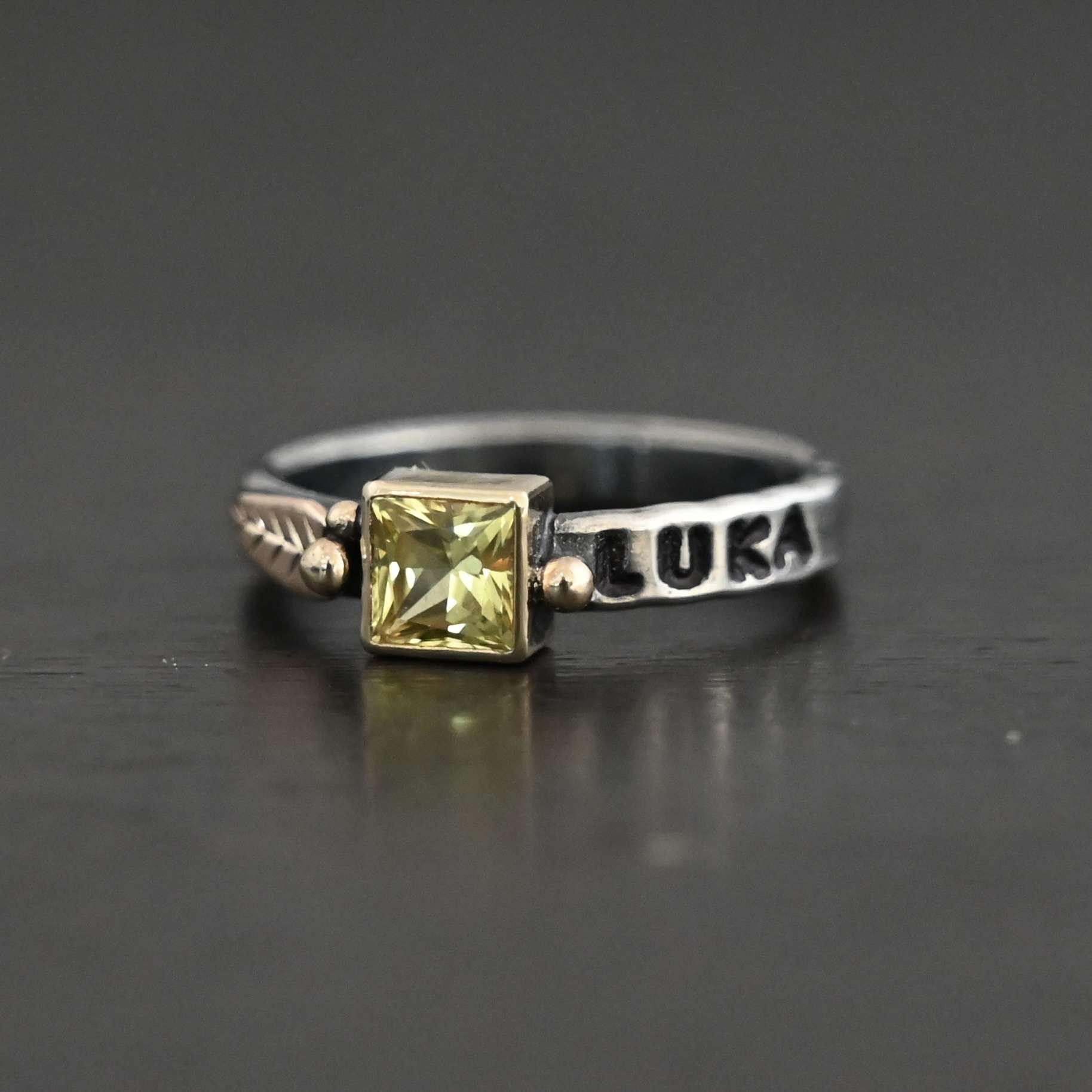Photo of a personalized birthstone stacker. Ring is a silver band with a square citrine in the center of it. The citrine is set in gold. There is a gold dot on the right side of the stone setting, and two on the left side. There is also a gold leaf on the left side of the stone setting. The name "Luka" is stamped and colored in with black on the right side of the ring. Ring is sitting against a black background.