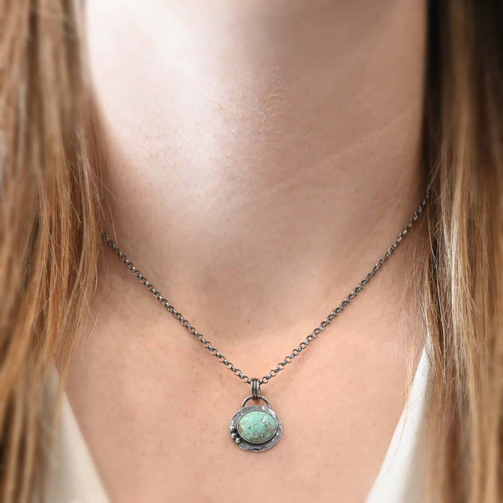 Close up of the "Forget Me Not" Necklace. Made out of turquoise and sterling silver. The turquoise is an oval shape and is a teal color with brown lines that are thick and thin squiggling all throughout the necklace. There are three small silver dots on the left side of the turquoise. The turquoise is set in silver and has a silver disk circling around it. The disk is hammered and oxidized. There is a loop on the back of the pendant and the pendant is hanging on three silver jump rings. Pendant is hanging on a "Rachel Rolo" Chain that has small but thick circular links and is oxidized. Necklace is laying against a models neck at 18" long.