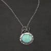 Close up of the "Forget Me Not" Necklace. Made out of turquoise and sterling silver. The turquoise is an oval shape and is a teal color with brown lines that are thick and thin squiggling all throughout the necklace. There are three small silver dots on the left side of the turquoise. The turquoise is set in silver and has a silver disk circling around it. The disk is hammered and oxidized. There is a loop on the back of the pendant and the pendant is hanging on three silver jump rings. Pendant is hanging on a "Rachel Rolo" Chain that has small but thick circular links and is oxidized. Necklace is laying on a dark background.