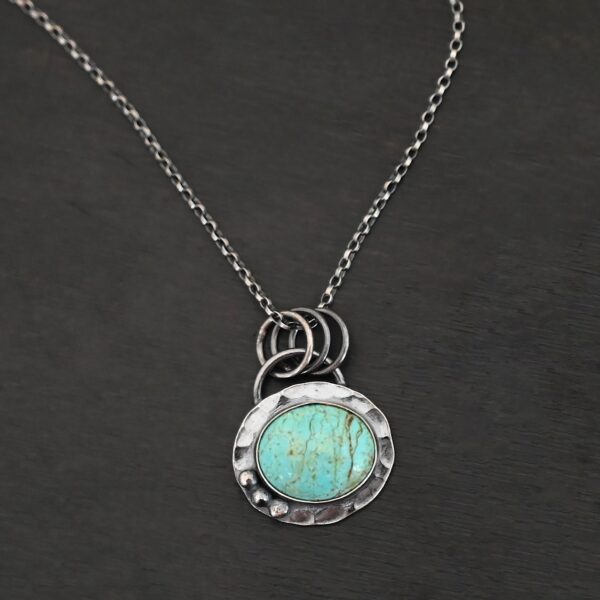 Close up of the "Forget Me Not" Necklace. Made out of turquoise and sterling silver. The turquoise is an oval shape and is a teal color with brown lines that are thick and thin squiggling all throughout the necklace. There are three small silver dots on the left side of the turquoise. The turquoise is set in silver and has a silver disk circling around it. The disk is hammered and oxidized. There is a loop on the back of the pendant and the pendant is hanging on three silver jump rings. Pendant is hanging on a "Rachel Rolo" Chain that has small but thick circular links and is oxidized. Necklace is laying on a dark background.