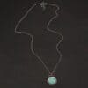 Close up of the "Forget Me Not" Necklace. Made out of turquoise and sterling silver. The turquoise is an oval shape and is a teal color with brown lines that are thick and thin squiggling all throughout the necklace. There are three small silver dots on the left side of the turquoise. The turquoise is set in silver and has a silver disk circling around it. The disk is hammered and oxidized. There is a loop on the back of the pendant and the pendant is hanging on three silver jump rings. Pendant is hanging on a "Rachel Rolo" Chain that has small but thick circular links and is oxidized. Necklace is laying on a dark background.