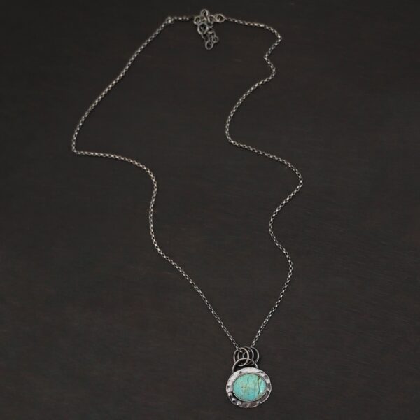 Close up of the "Forget Me Not" Necklace. Made out of turquoise and sterling silver. The turquoise is an oval shape and is a teal color with brown lines that are thick and thin squiggling all throughout the necklace. There are three small silver dots on the left side of the turquoise. The turquoise is set in silver and has a silver disk circling around it. The disk is hammered and oxidized. There is a loop on the back of the pendant and the pendant is hanging on three silver jump rings. Pendant is hanging on a "Rachel Rolo" Chain that has small but thick circular links and is oxidized. Necklace is laying on a dark background.