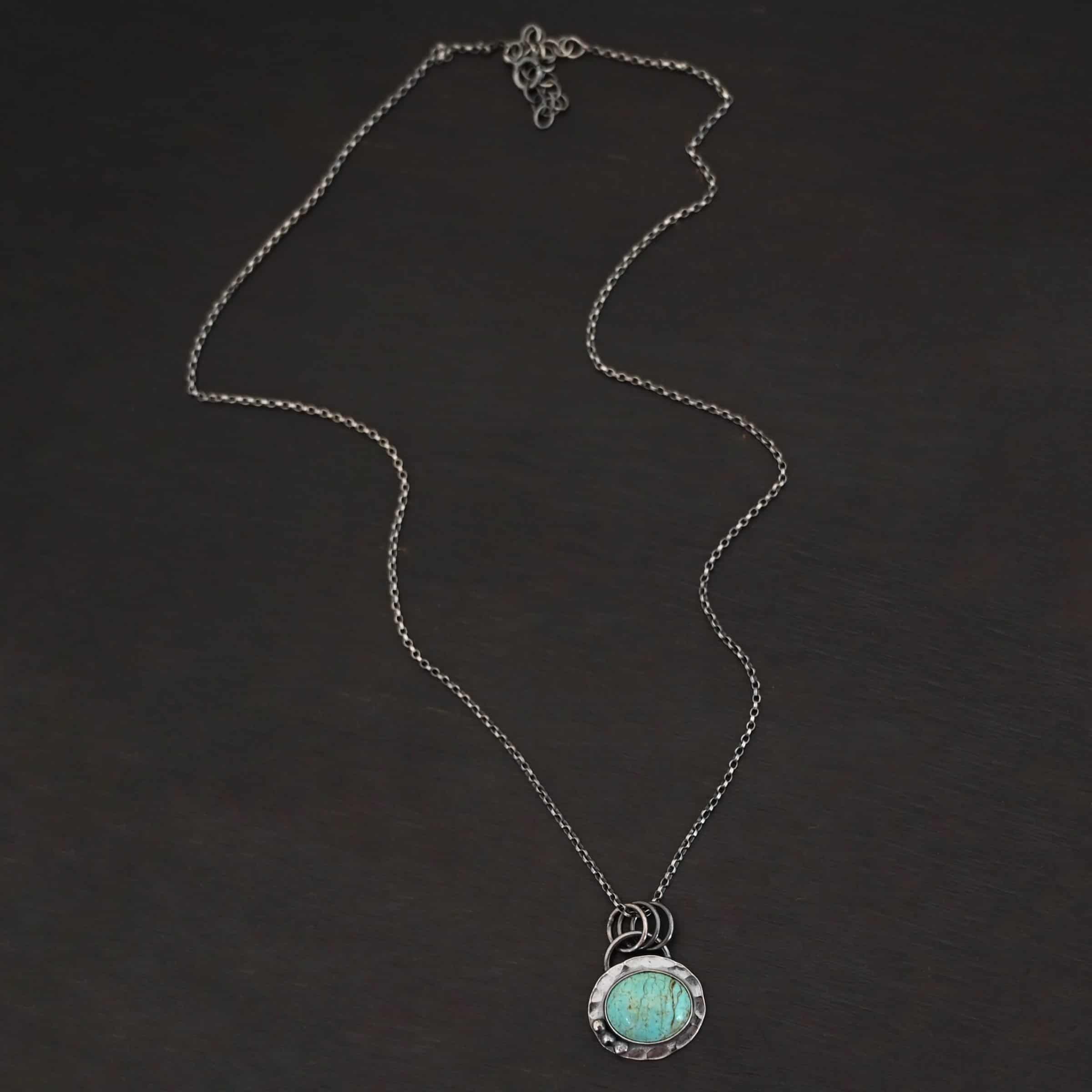 Close up of the "Forget Me Not" Necklace. Made out of turquoise and sterling silver. The turquoise is an oval shape and is a teal color with brown lines that are thick and thin squiggling all throughout the necklace. There are three small silver dots on the left side of the turquoise. The turquoise is set in silver and has a silver disk circling around it. The disk is hammered and oxidized. There is a loop on the back of the pendant and the pendant is hanging on three silver jump rings. Pendant is hanging on a "Rachel Rolo" Chain that has small but thick circular links and is oxidized. Necklace is laying on a dark background.