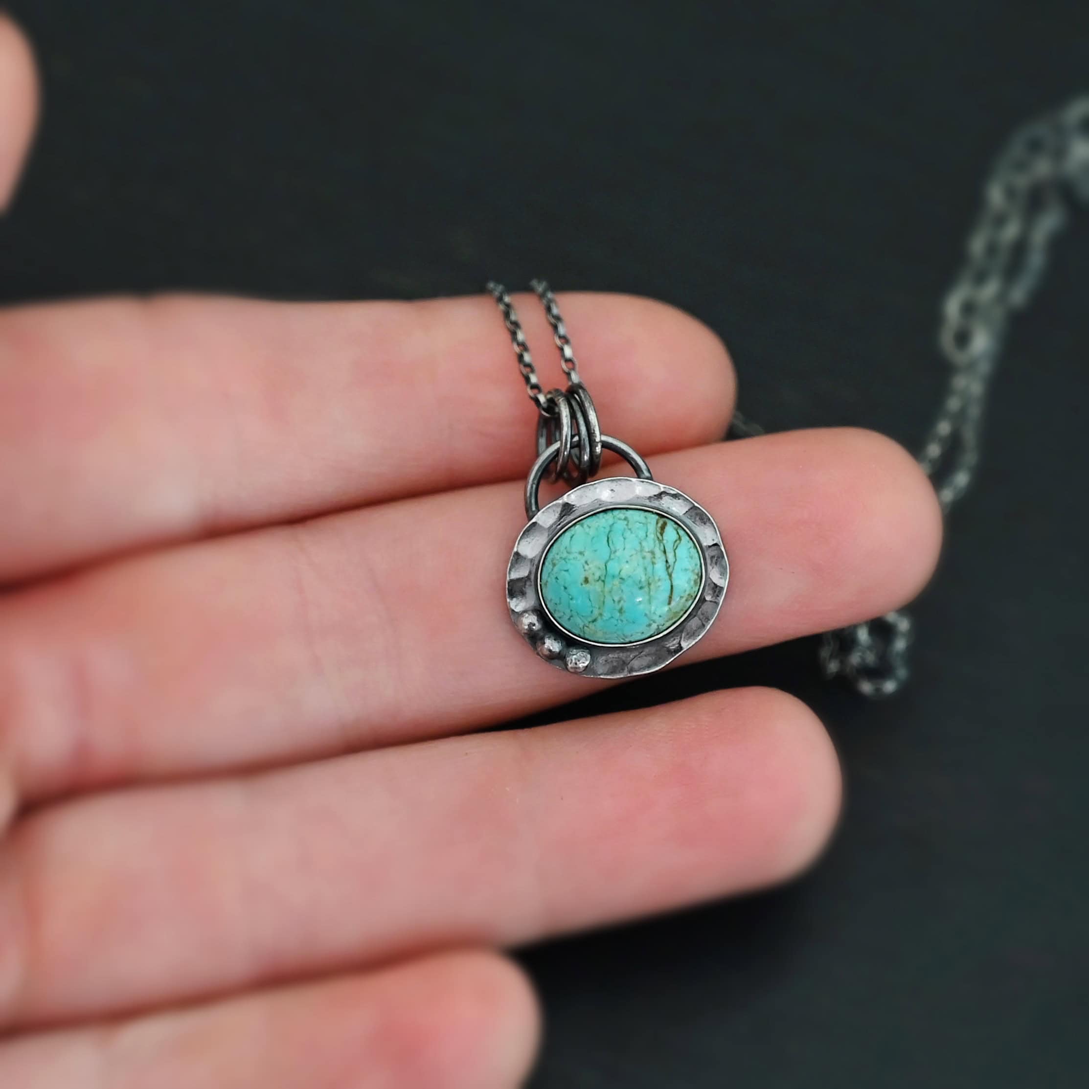 Close up of the "Forget Me Not" Necklace. Made out of turquoise and sterling silver. The turquoise is an oval shape and is a teal color with brown lines that are thick and thin squiggling all throughout the necklace. There are three small silver dots on the left side of the turquoise. The turquoise is set in silver and has a silver disk circling around it. The disk is hammered and oxidized. There is a loop on the back of the pendant and the pendant is hanging on three silver jump rings. Pendant is hanging on a "Rachel Rolo" Chain that has small but thick circular links and is oxidized. Necklace is laying on the fingertips of a hand to demonstrate size of the pendant.