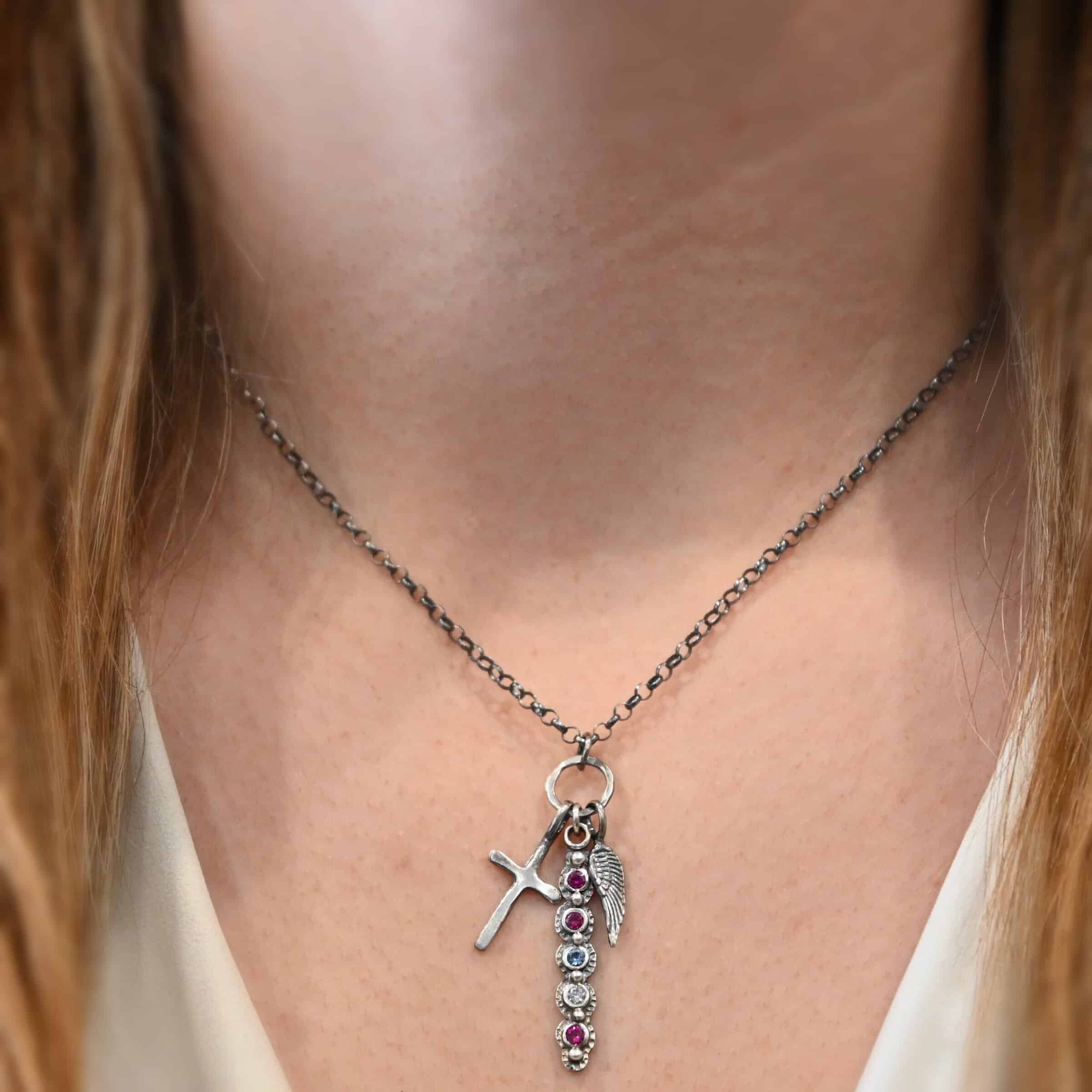 Photo of a "Mother's Necklace" that was custom. Made out of sterling silver and five different birthstones. They go in a straight line from top to bottom. The first two towards the top are pink colored. Then there is a light blue color. Then a clear stone. Finally, another pink stone at the end. Each stone is a small circle, and the silver around them is hammered and oxidized. There are six silver balls on the necklace that go in a straight line. There is one on the top of the first stone, and each ball after that separates each stone. There is a loop at the top of the pendant with a jump ring through it that is attached to a larger hammered circle. There is a small cross charm on the left side of the pendant, and an angel wing charm on the right side of the pendant. Both are attached to the large circle as well. The large circle has a small jump ring attached to the top of it in which the chain goes through. Necklace is laying against a model's neck to demonstrate size. Necklace is worn at 17 inches long, and model is wearing a white v-neck shirt.