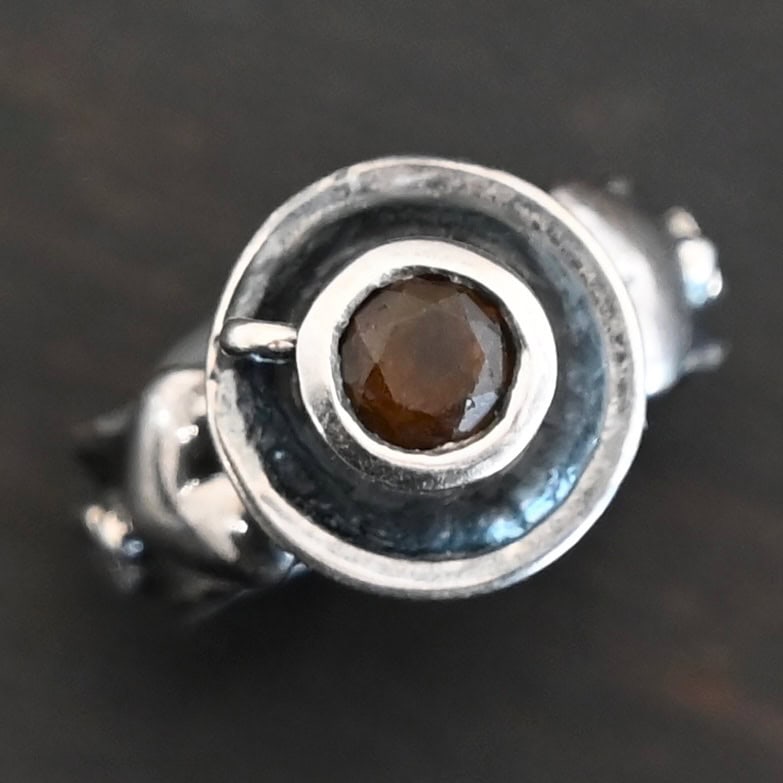 True Addiction Coffee Ring "True Addiction Coffee" Ring as shown up close. Made out of sterling silver and topaz. Small sterling silver coffee cup with the topaz in the center to look as if it is filled with coffee. Coffee cup sits on a small sterling silver disk to look as if it is sitting on a plate. There are two hearts on the ring, one on the left side and one on the right. Ring sits against a dark background.