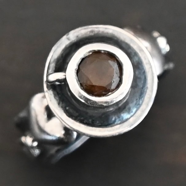 True Addiction Coffee Ring "True Addiction Coffee" Ring as shown up close. Made out of sterling silver and topaz. Small sterling silver coffee cup with the topaz in the center to look as if it is filled with coffee. Coffee cup sits on a small sterling silver disk to look as if it is sitting on a plate. There are two hearts on the ring, one on the left side and one on the right. Ring sits against a dark background.