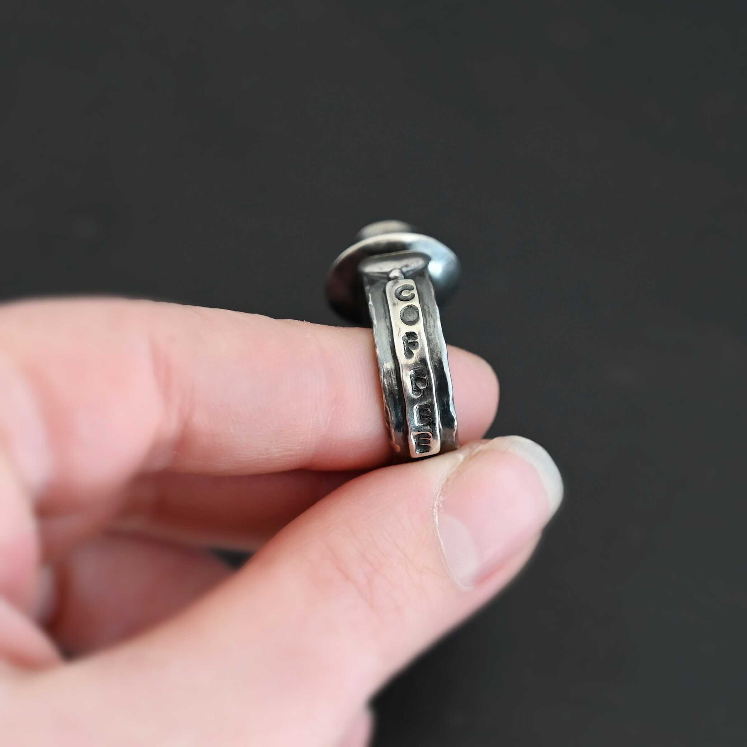 True Addiction Coffee Ring "True Addiction Coffee" Ring as shown up close. Made out of sterling silver and topaz. Small sterling silver coffee cup with the topaz in the center to look as if it is filled with coffee. Coffee cup sits on a small sterling silver disk to look as if it is sitting on a plate. There are two hearts on the ring, one on the left side and one on the right. Ring is held in the fingertips of a hand to show the word "coffee" stamped vertically into the ring down one side of it.