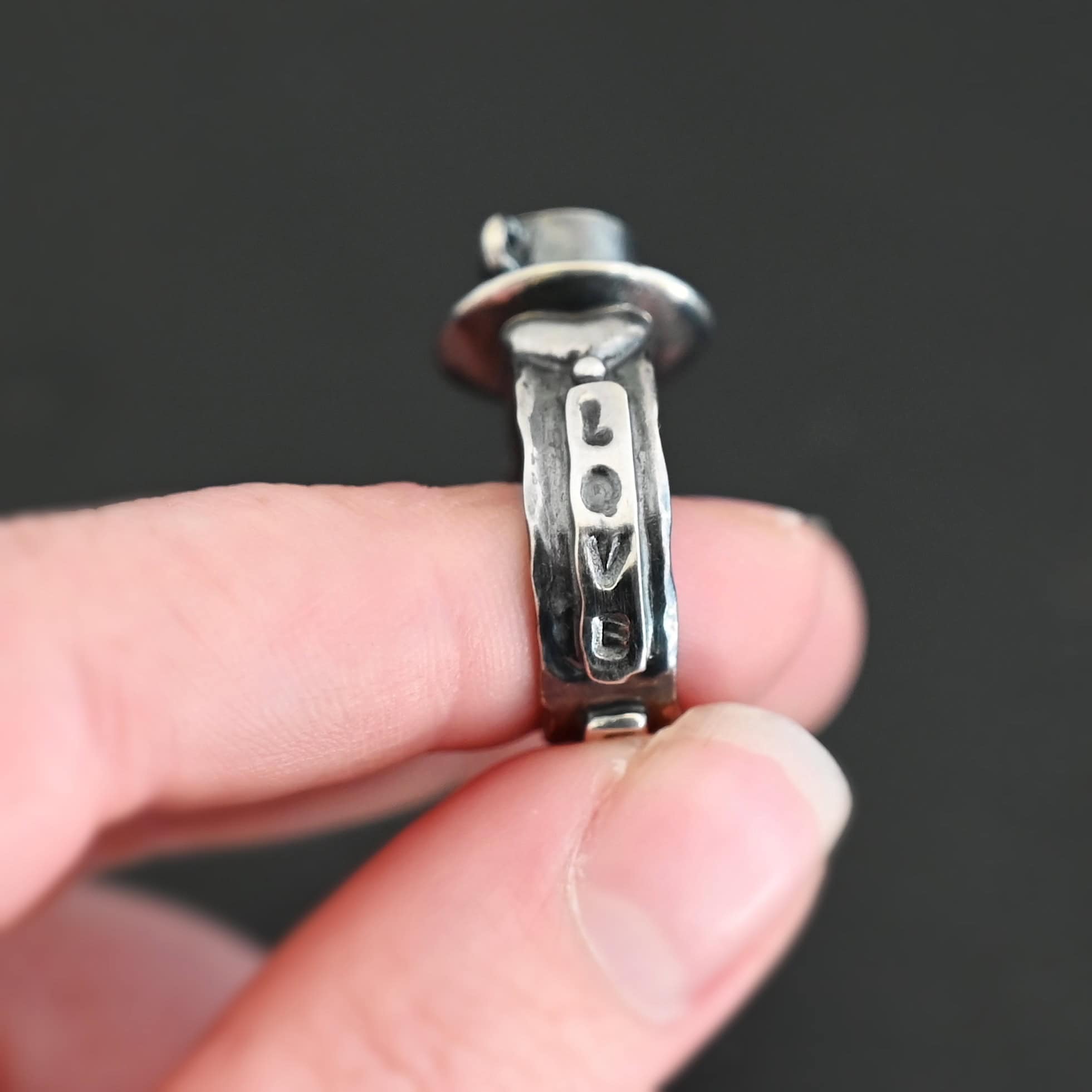 True Addiction Coffee Ring "True Addiction Coffee" Ring as shown up close. Made out of sterling silver and topaz. Small sterling silver coffee cup with the topaz in the center to look as if it is filled with coffee. Coffee cup sits on a small sterling silver disk to look as if it is sitting on a plate. There are two hearts on the ring, one on the left side and one on the right. Ring is held in the fingertips of a hand to show the word "love" stamped vertically into the ring down one side of it.