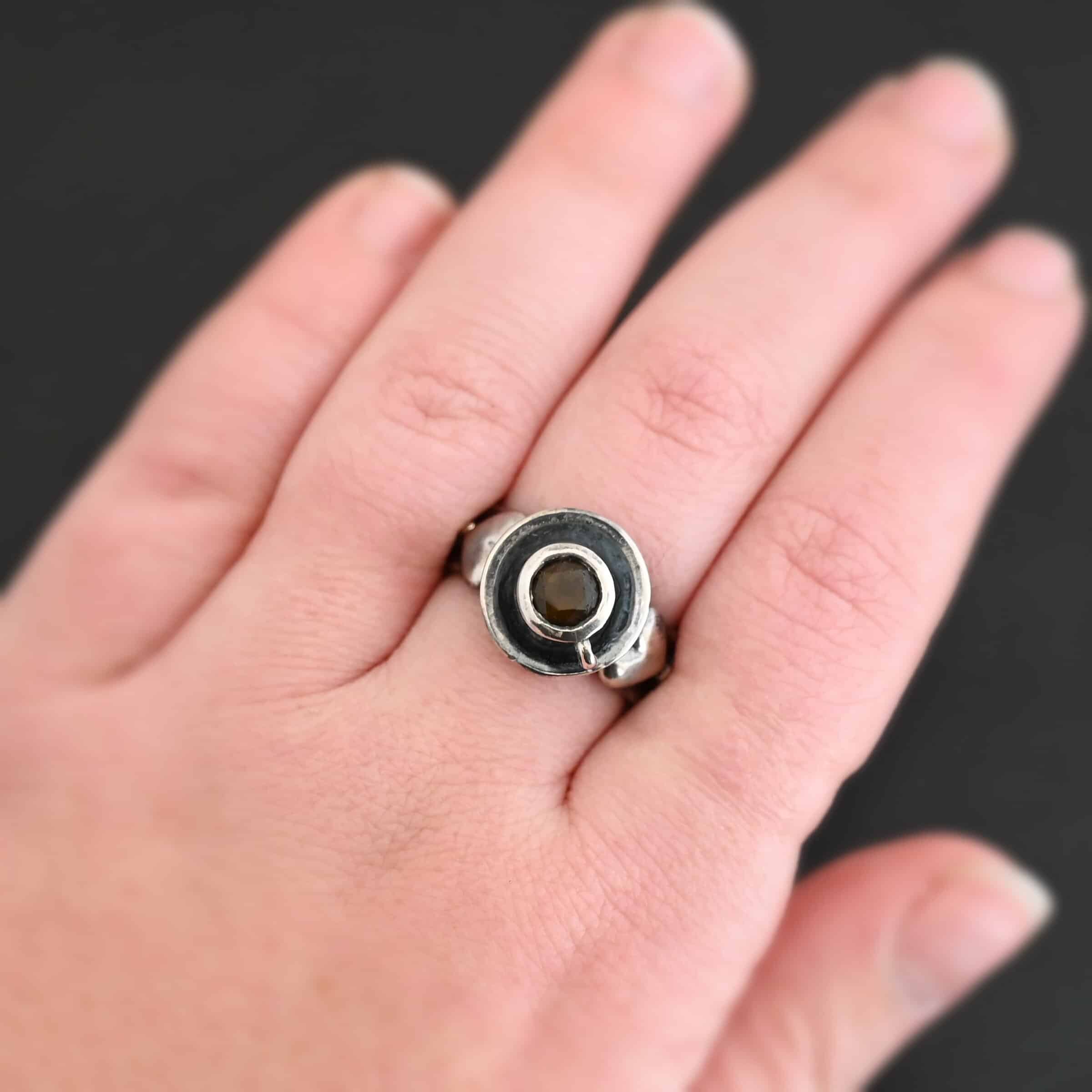 True Addiction Coffee Ring "True Addiction Coffee" Ring as shown up close. Made out of sterling silver and topaz. Small sterling silver coffee cup with the topaz in the center to look as if it is filled with coffee. Coffee cup sits on a small sterling silver disk to look as if it is sitting on a plate. There are two hearts on the ring, one on the left side and one on the right. Ring is worn by a model up close.