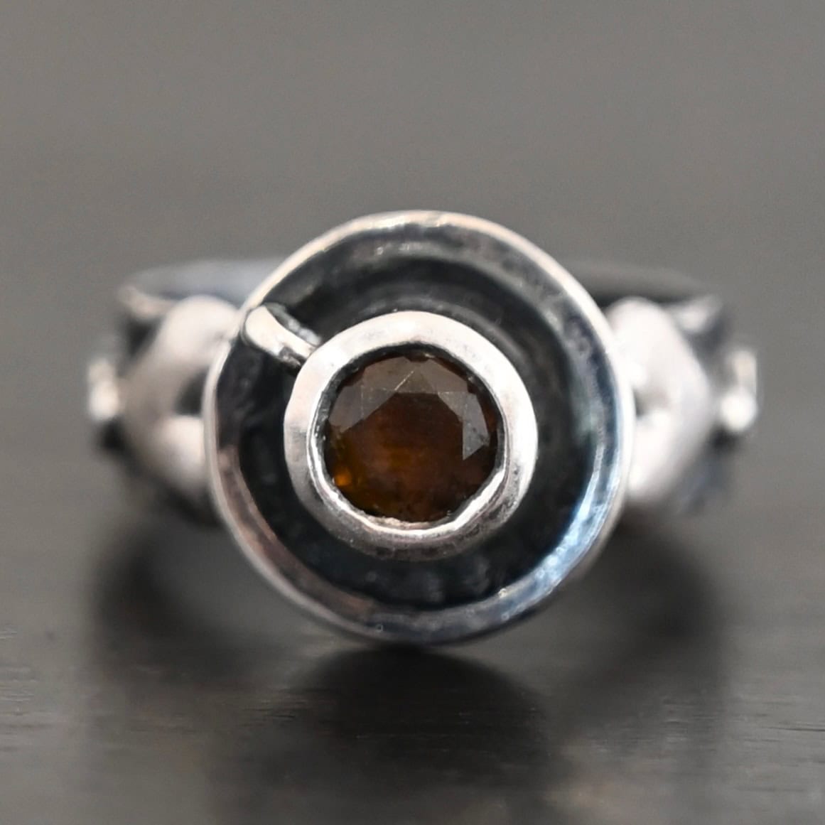 True Addiction Coffee Ring "True Addiction Coffee" Ring as shown up close. Made out of sterling silver and topaz. Small sterling silver coffee cup with the topaz in the center to look as if it is filled with coffee. Coffee cup sits on a small sterling silver disk to look as if it is sitting on a plate. There are two hearts on the ring, one on the left side and one on the right. Ring sits against a dark background.