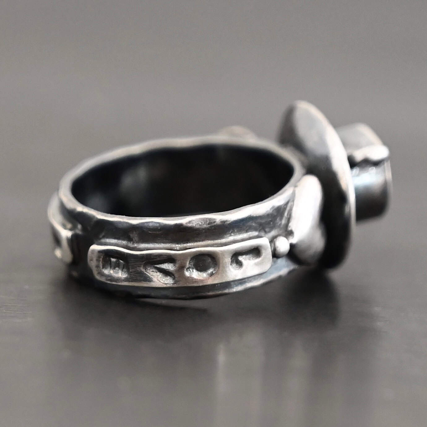 True Addiction Coffee Ring "True Addiction Coffee" Ring as shown up close. Made out of sterling silver and topaz. Small sterling silver coffee cup with the topaz in the center to look as if it is filled with coffee. Coffee cup sits on a small sterling silver disk to look as if it is sitting on a plate. There are two hearts on the ring, one on the left side and one on the right. Ring sits against a dark background.