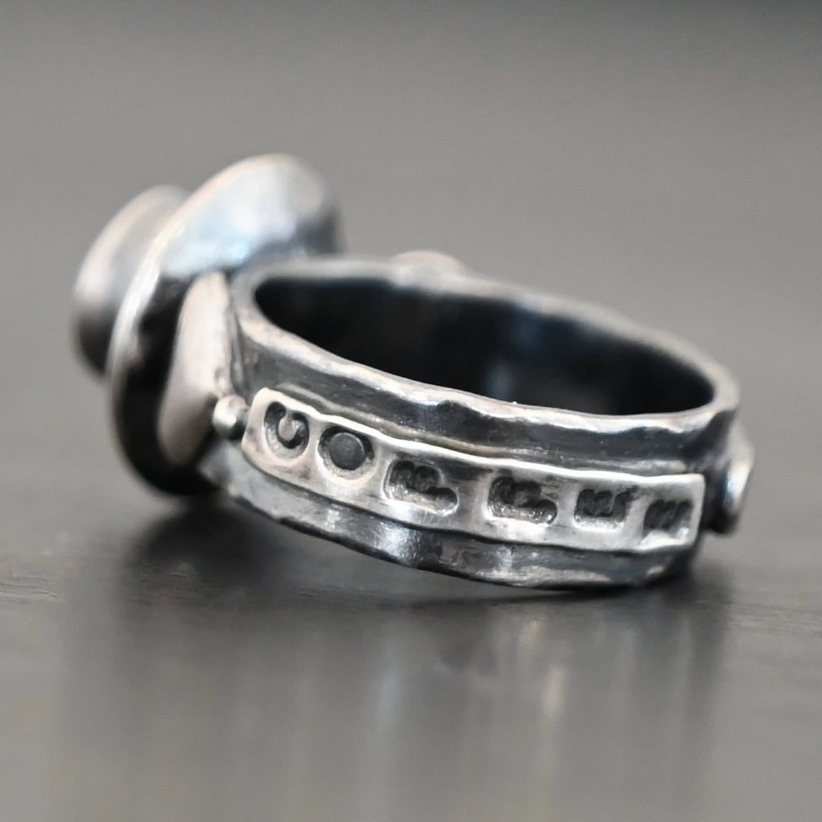 True Addiction Coffee Ring "True Addiction Coffee" Ring as shown up close. Made out of sterling silver and topaz. Small sterling silver coffee cup with the topaz in the center to look as if it is filled with coffee. Coffee cup sits on a small sterling silver disk to look as if it is sitting on a plate. There are two hearts on the ring, one on the left side and one on the right. Ring sits against a dark background.