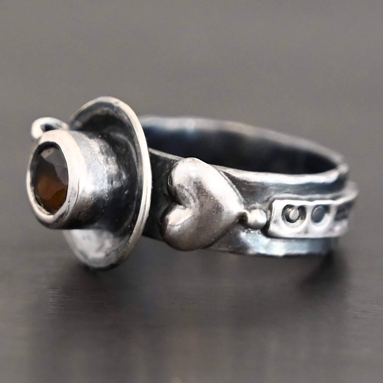 True Addiction Coffee Ring "True Addiction Coffee" Ring as shown up close. Made out of sterling silver and topaz. Small sterling silver coffee cup with the topaz in the center to look as if it is filled with coffee. Coffee cup sits on a small sterling silver disk to look as if it is sitting on a plate. There are two hearts on the ring, one on the left side and one on the right. Ring sits against a dark background.