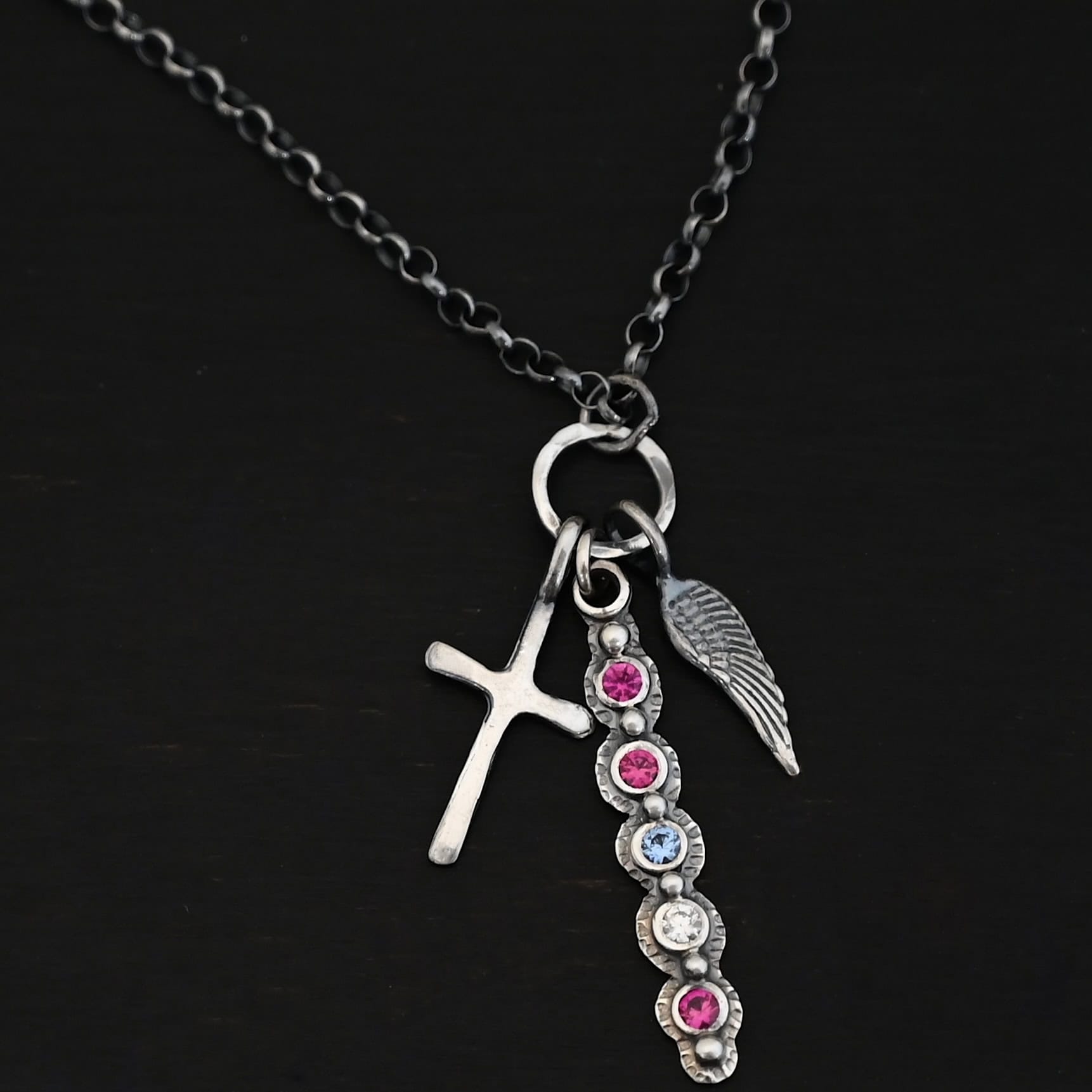 Photo of a "Mother's Necklace" that was custom. Made out of sterling silver and five different birthstones. They go in a straight line from top to bottom. The first two towards the top are pink colored. Then there is a light blue color. Then a clear stone. Finally, another pink stone at the end. Each stone is a small circle, and the silver around them is hammered and oxidized. There are six silver balls on the necklace that go in a straight line. There is one on the top of the first stone, and each ball after that separates each stone. There is a loop at the top of the pendant with a jump ring through it that is attached to a larger hammered circle. There is a small cross charm on the left side of the pendant, and an angel wing charm on the right side of the pendant. Both are attached to the large circle as well. The large circle has a small jump ring attached to the top of it in which the chain goes through. Necklace is laying against a dark background.