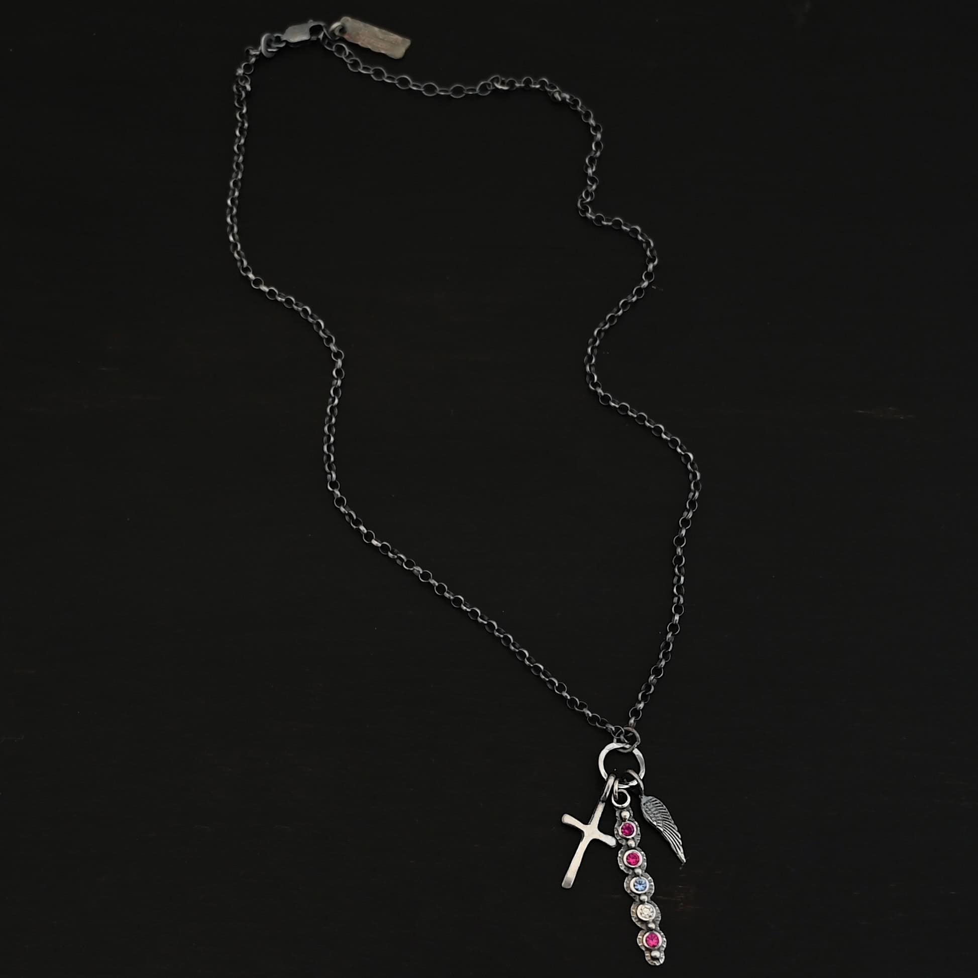 Photo of a "Mother's Necklace" that was custom. Made out of sterling silver and five different birthstones. They go in a straight line from top to bottom. The first two towards the top are pink colored. Then there is a light blue color. Then a clear stone. Finally, another pink stone at the end. Each stone is a small circle, and the silver around them is hammered and oxidized. There are six silver balls on the necklace that go in a straight line. There is one on the top of the first stone, and each ball after that separates each stone. There is a loop at the top of the pendant with a jump ring through it that is attached to a larger hammered circle. There is a small cross charm on the left side of the pendant, and an angel wing charm on the right side of the pendant. Both are attached to the large circle as well. The large circle has a small jump ring attached to the top of it in which the chain goes through. Necklace is laying against a dark background.