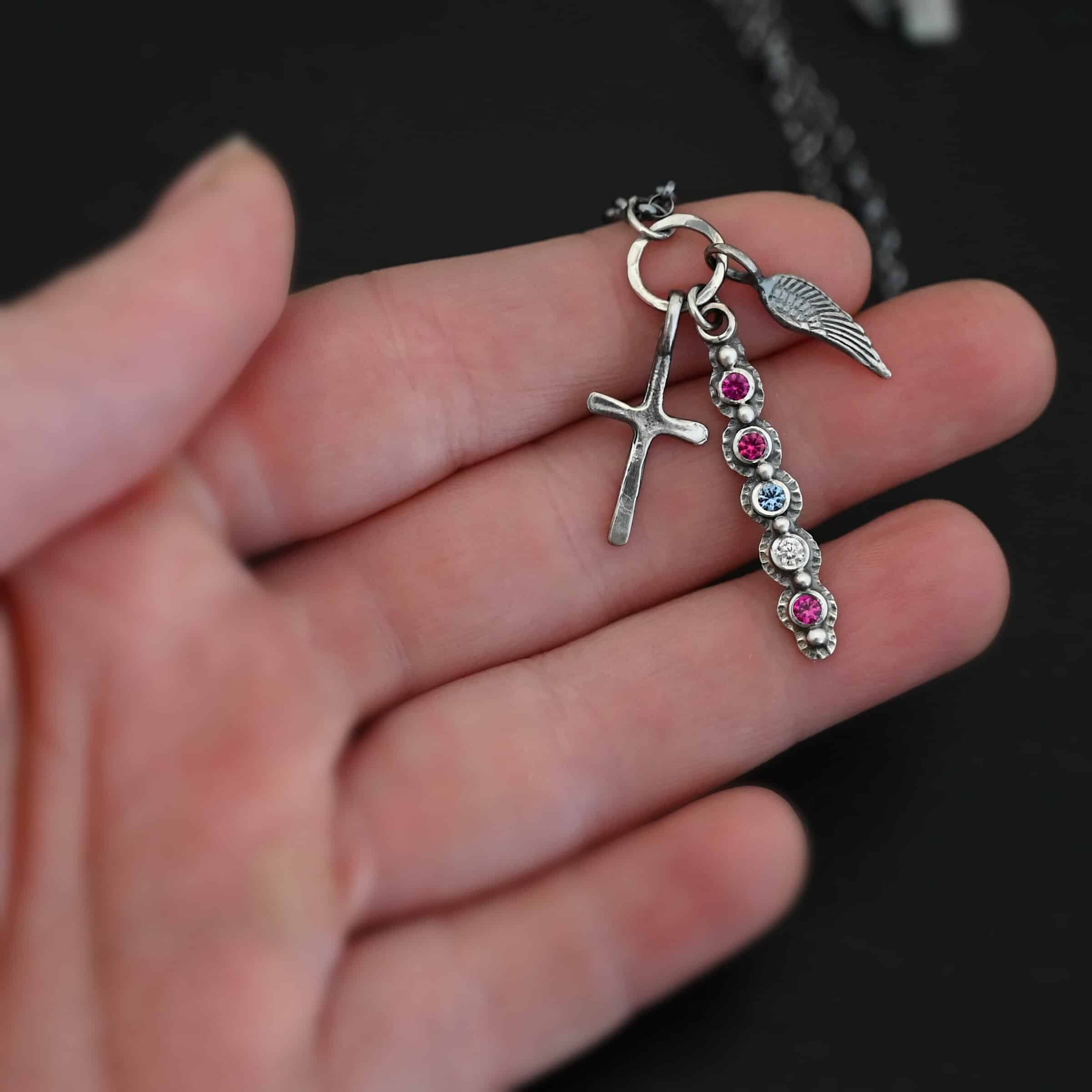 Photo of a "Mother's Necklace" that was custom. Made out of sterling silver and five different birthstones. They go in a straight line from top to bottom. The first two towards the top are pink colored. Then there is a light blue color. Then a clear stone. Finally, another pink stone at the end. Each stone is a small circle, and the silver around them is hammered and oxidized. There are six silver balls on the necklace that go in a straight line. There is one on the top of the first stone, and each ball after that separates each stone. There is a loop at the top of the pendant with a jump ring through it that is attached to a larger hammered circle. There is a small cross charm on the left side of the pendant, and an angel wing charm on the right side of the pendant. Both are attached to the large circle as well. The large circle has a small jump ring attached to the top of it in which the chain goes through. Necklace is laying against the fingertips of a hand to demonstrate size.