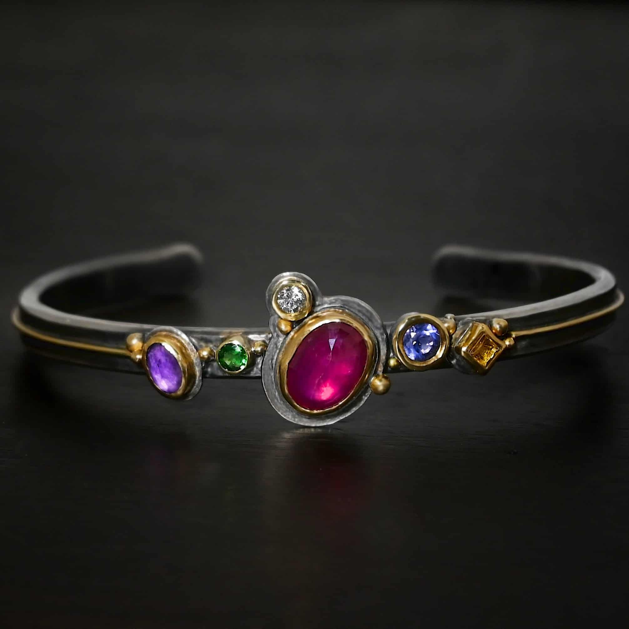 Cinderella Cuff Photo of the "Cinderella" Cuff. It has a ruby in the center, oval shaped and set sideways. There is an amethyst and tsavorite garnet on the left side and a citrine and amethyst on the right side. There is a diamond on the top left side of the ruby. Each stone is set in gold. There is a thin line of gold going across the cuff through the center on each side where the stones aren't. There are balls of gold scattered around the stones. Cuff is lying against a dark background.